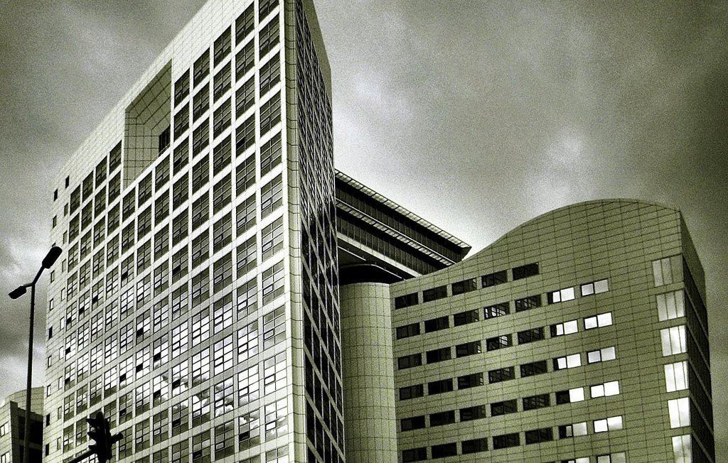 L'image montre un bâtiment moderne avec une architecture contemporaine. La structure est composée de plusieurs angles et surfaces réfléchissantes, donnant une impression de sophistication. Les vitres sont grandes, et le design présente des lignes épurées. Le ciel en arrière-plan est nuageux, ce qui peut créer une ambiance dramatique. Les tons de l'image sont en noir et blanc, accentuant les contrastes entre la lumière et l'ombre sur la façade du bâtiment.