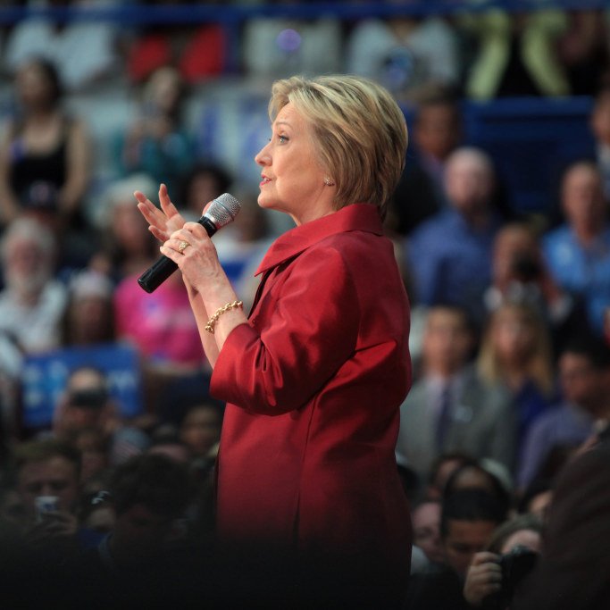 L'image montre une femme prenant la parole devant un public. Elle porte un manteau rouge et utilise un microphone pour s'exprimer. En arrière-plan, on peut voir une foule de personnes, certaines tenant des panneaux ou des affiches. L'éclairage et l'ambiance suggèrent un événement politique ou une réunion publique.