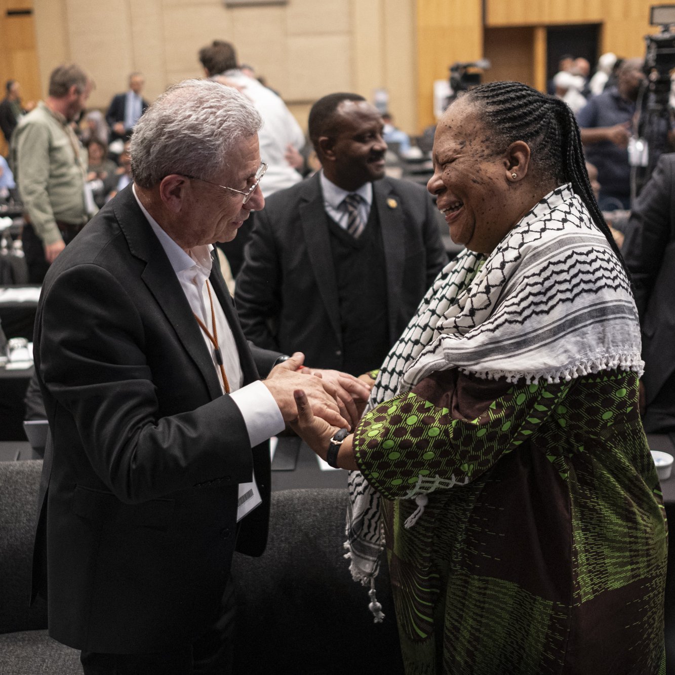 L'image montre deux personnes en train d'échanger des sourires et de se serrer la main dans un environnement formel, probablement lors d'une conférence ou d'un événement. Ils semblent engagés dans une conversation amicale, entourés de plusieurs autres personnes en arrière-plan. L'ambiance paraît chaleureuse et conviviale, avec un focus sur l'interaction entre ces deux individus. Les participants portent des vêtements variés, ajoutant à la diversité de la scène.