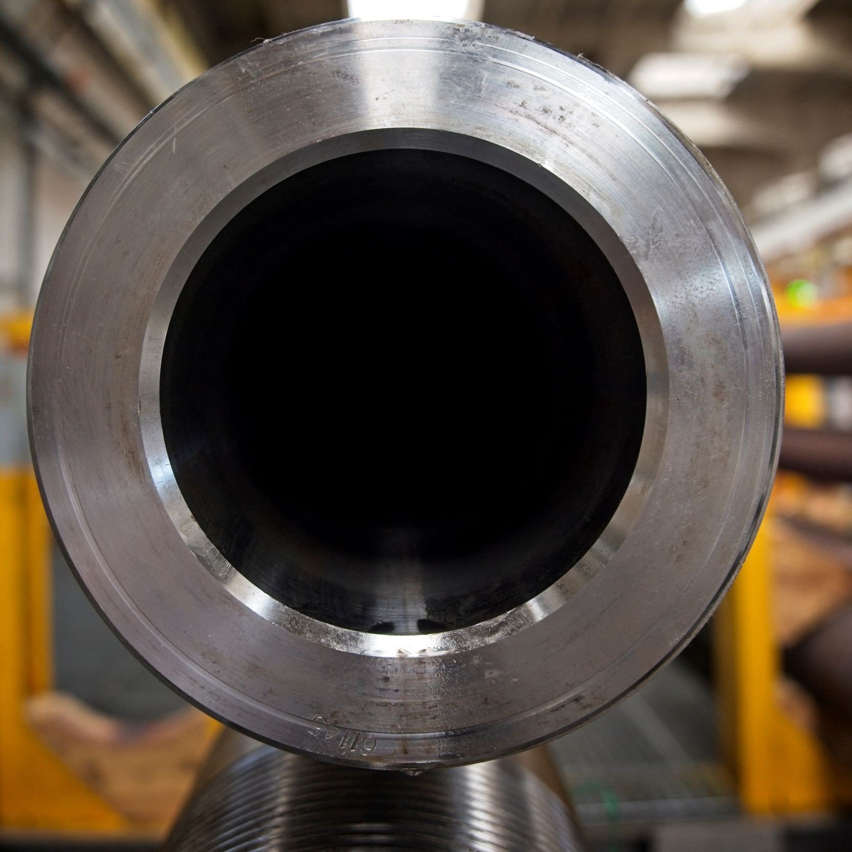 L'image montre la vue de l'intérieur d'un grand tube métallique. On peut voir la texture lisse et brillante du métal au niveau de l'ouverture, tandis que l'intérieur du tube est sombre. En arrière-plan, des éléments d'un environnement industriel sont visibles, avec d'autres tubes et structures, suggérant un lieu de fabrication ou d'assemblage. L'éclairage est principalement naturel, créant un effet contrasté sur la surface du tube.