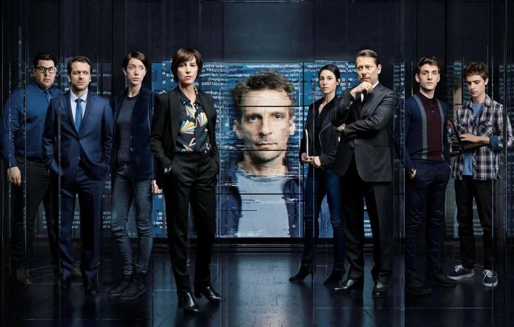 L'image présente un groupe de personnes stylisées, probablement des acteurs, se tenant debout dans un environnement sombre et futuriste. Au centre, un écran numérique affiche le visage d'un homme, tandis que les membres du groupe adoptent des poses sérieuses. Ils portent des vêtements formels et décontractés, suggérant une ambiance à la fois professionnelle et tendue. L'ensemble de l'image évoque un thème lié à la technologie ou aux enquêtes, avec une atmosphère de mystère.