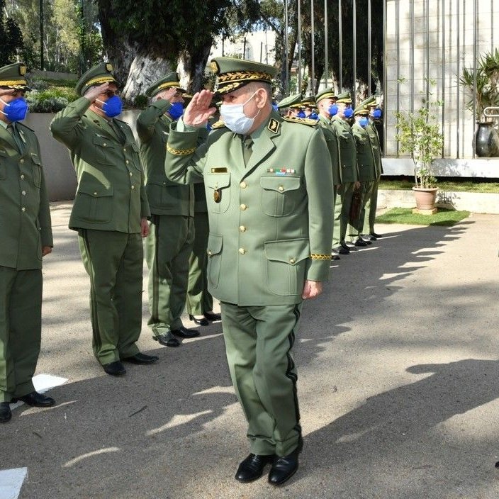 L'image montre un groupe de militaires en uniforme, saluant un supérieur. Tous portent des masques et des casquettes, ce qui indique qu'ils respectent des protocoles de santé. L'environnement semble être un site officiel ou militaire, avec des bâtiments en arrière-plan. Les militaires adoptent une posture de respect, ce qui suggère une cérémonie ou un événement formel.