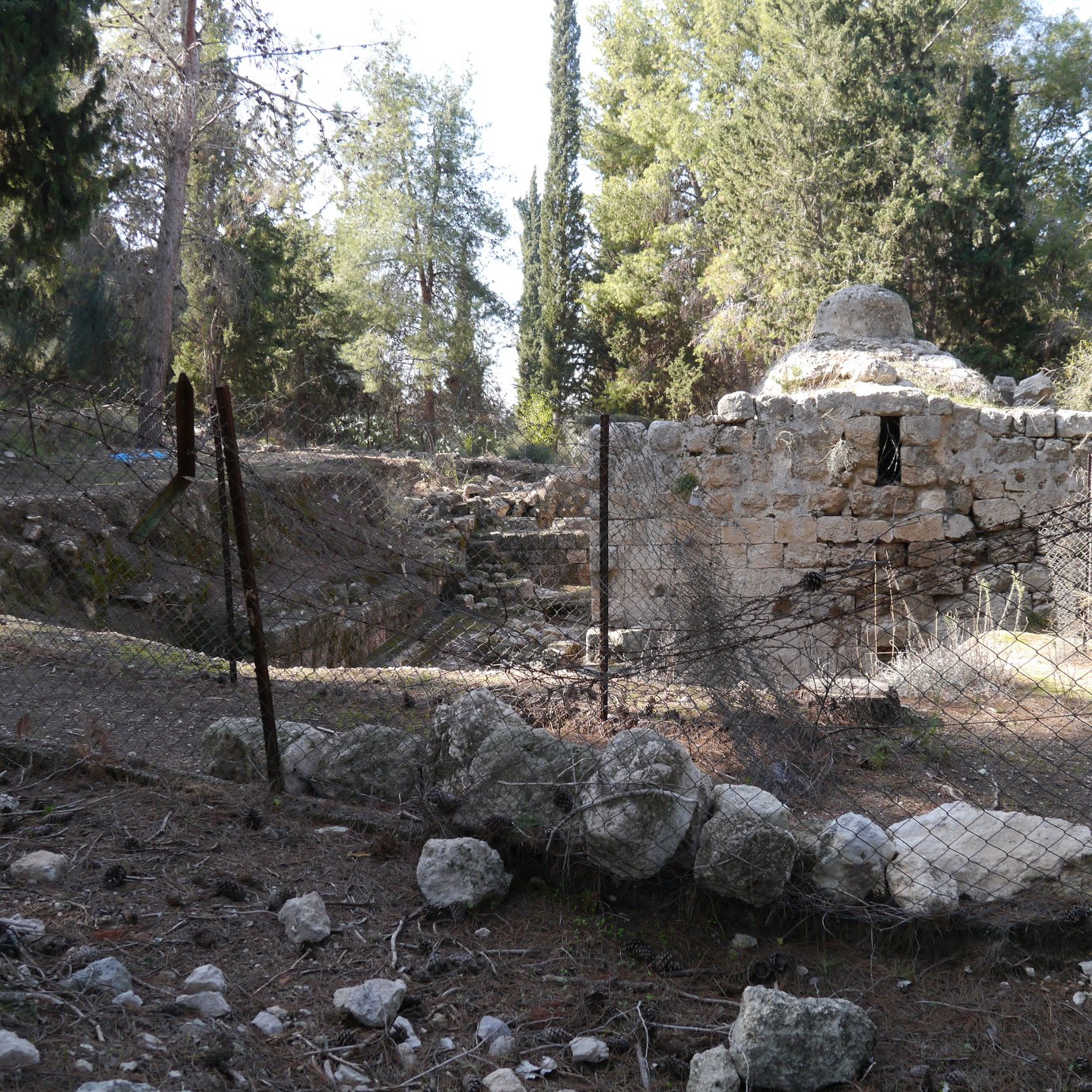 L'image montre un site archéologique entouré d'une clôture en fil de fer. Il y a des ruines anciennes, probablement d'une structure en pierre, avec des éléments végétaux environnants tels que des arbres et des buissons. Le sol est rempli de pierres et de débris, ce qui donne une impression d'abandon. L'atmosphère semble paisible, mais aussi légèrement mélancolique, témoignant d'un passé oublié.