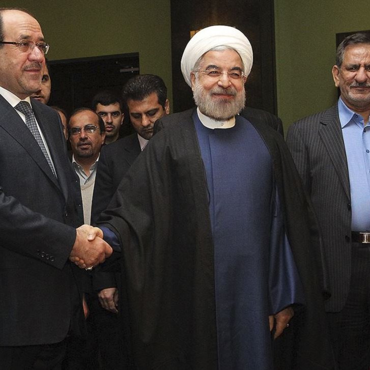 L'image montre trois hommes se tenant debout, avec un homme au centre qui sourit et porte une robe traditionnelle, tandis que les deux autres sont en costume. Ils semblent être en train de se saluer, probablement lors d'une rencontre officielle. L'environnement semble formel, probablement dans un hôtel ou un bâtiment gouvernemental, avec des personnes en arrière-plan.