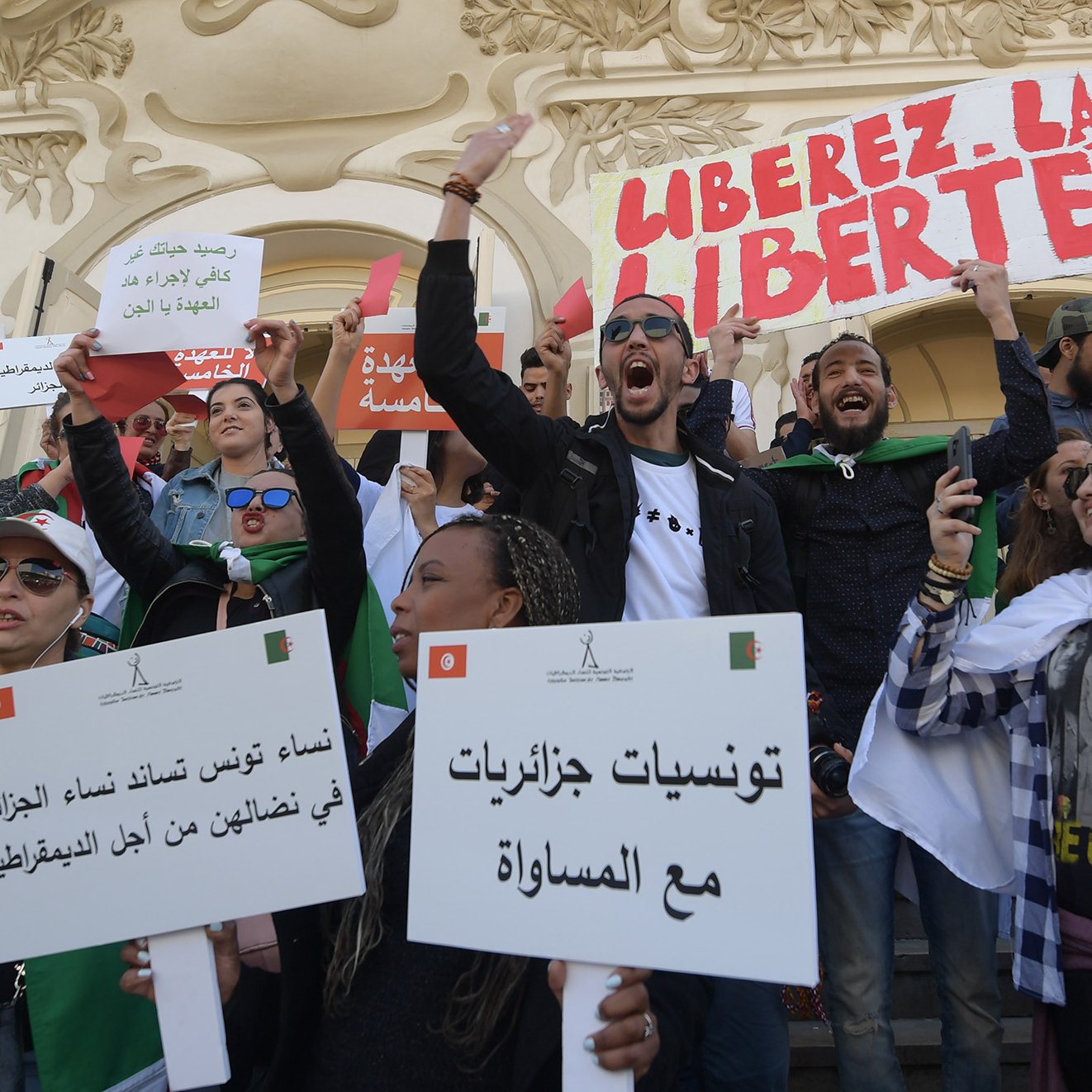 L'image montre un groupe de personnes rassemblées lors d'une manifestation. Les manifestants portent des pancartes et des drapeaux, exprimant des slogans tels que "Libérez la liberté". Les participants semblent engagés et passionnés, brandissant des affiches avec des messages en arabe et en français, appelant à la justice et à l'égalité. L'ambiance est énergique et déterminée, témoignant d'une dynamique de revendication sociale ou politique. L'arrière-plan présente une architecture caractéristique, ajoutant au contexte de l'événement.