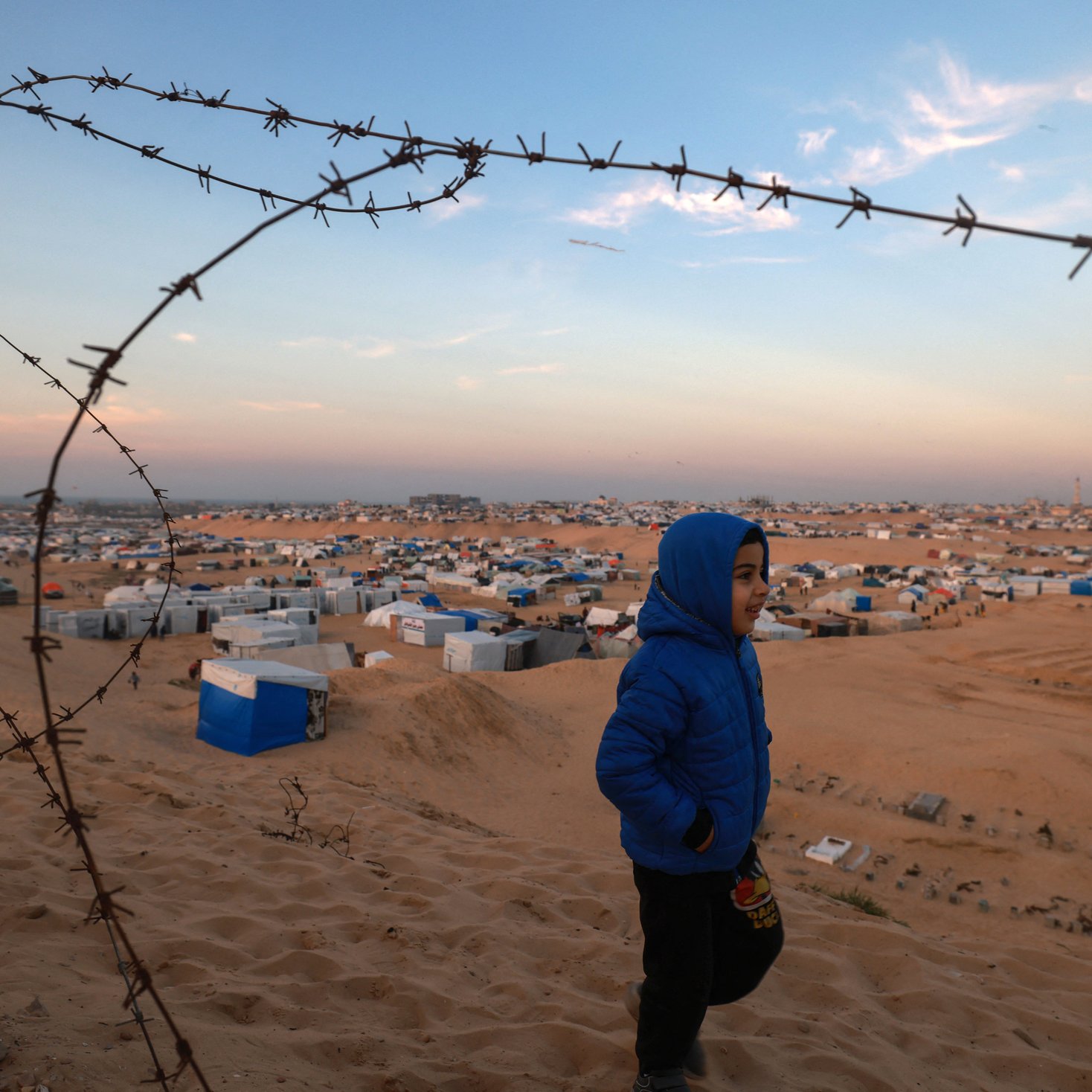 The image shows a child wearing a blue jacket walking along a sandy landscape. In the background, there are numerous temporary shelters, likely tents or makeshift homes, scattered across the terrain. The sky is a mix of soft colors, indicating either dawn or dusk. A fence topped with barbed wire runs along the foreground, adding a sense of restriction or boundary to the scene. The overall atmosphere conveys a sense of hardship, amidst the child's movement.