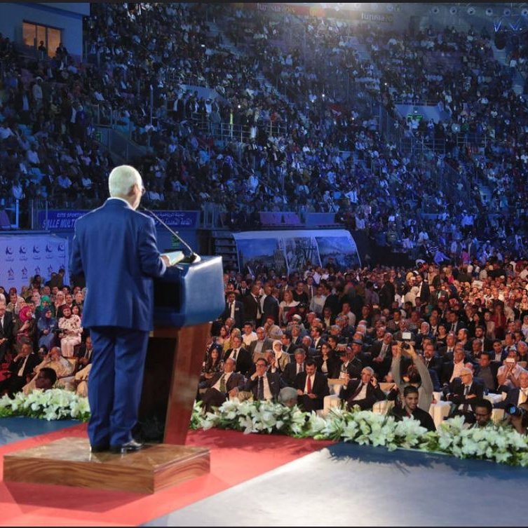 L'image montre un homme debout sur une scène, s'adressant à une foule nombreuse. Il est en costume et se tient derrière un pupitre, probablement en train de donner un discours. La salle est pleine, avec des centaines de personnes assises en face de lui, certaines regardant attentivement. L'éclairage crée une ambiance dramatique, mettant en valeur à la fois le conférencier et le public. Des décorations florales sont visibles au premier plan, ajoutant une touche d'élégance à l'événement.