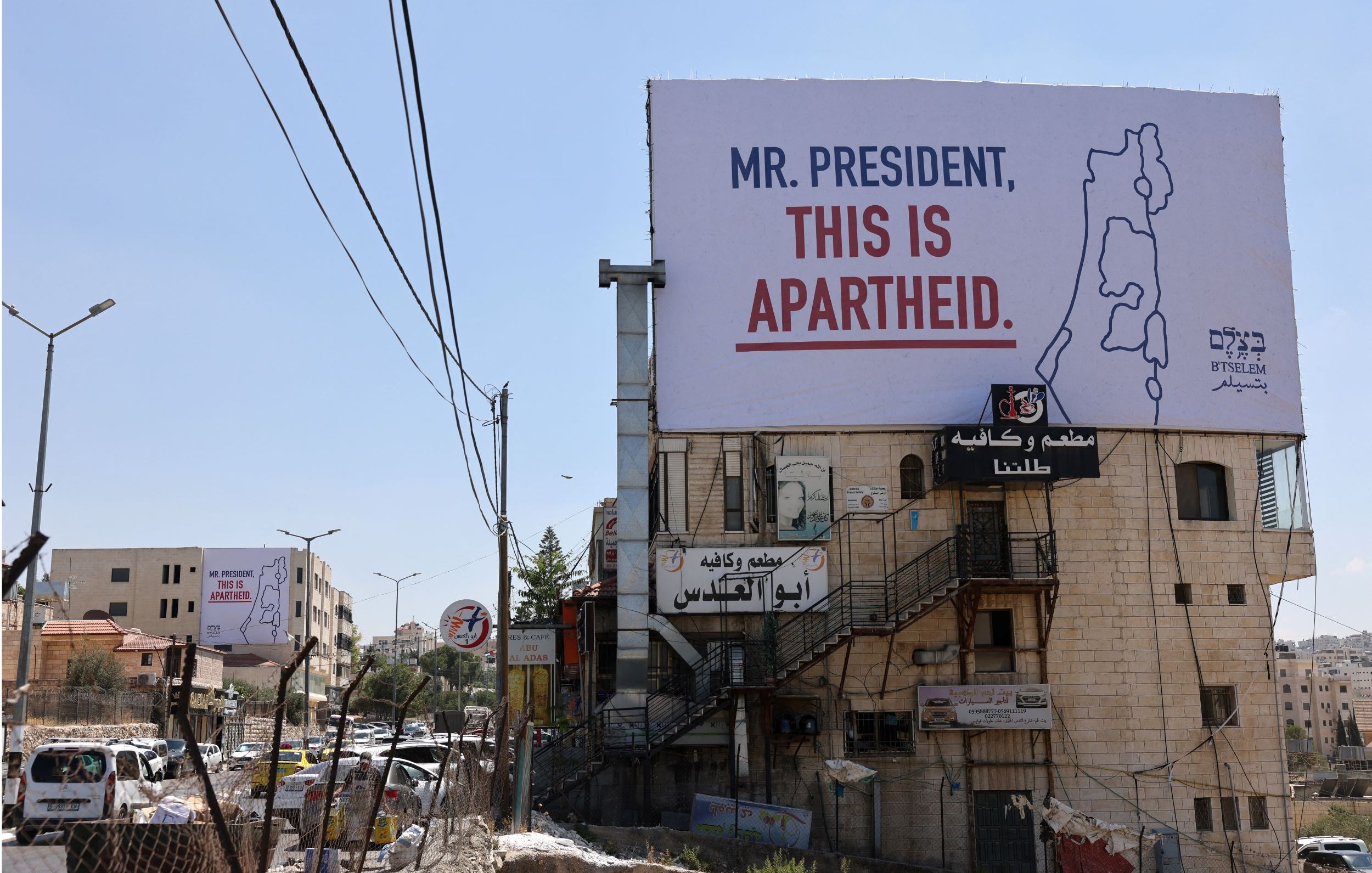 L'image montre un panneau publicitaire accroché sur un bâtiment, avec un message qui dit "MR. PRESIDENT, THIS IS APARTHEID." On peut voir des infrastructures urbaines environnantes, telles que des routes, des bâtiments et des fils de téléphone. Le contexte suggère une critique sociale ou politique liée à la situation en Israël-Palestine. Le paysage est ensoleillé et présente une ambiance de ville.