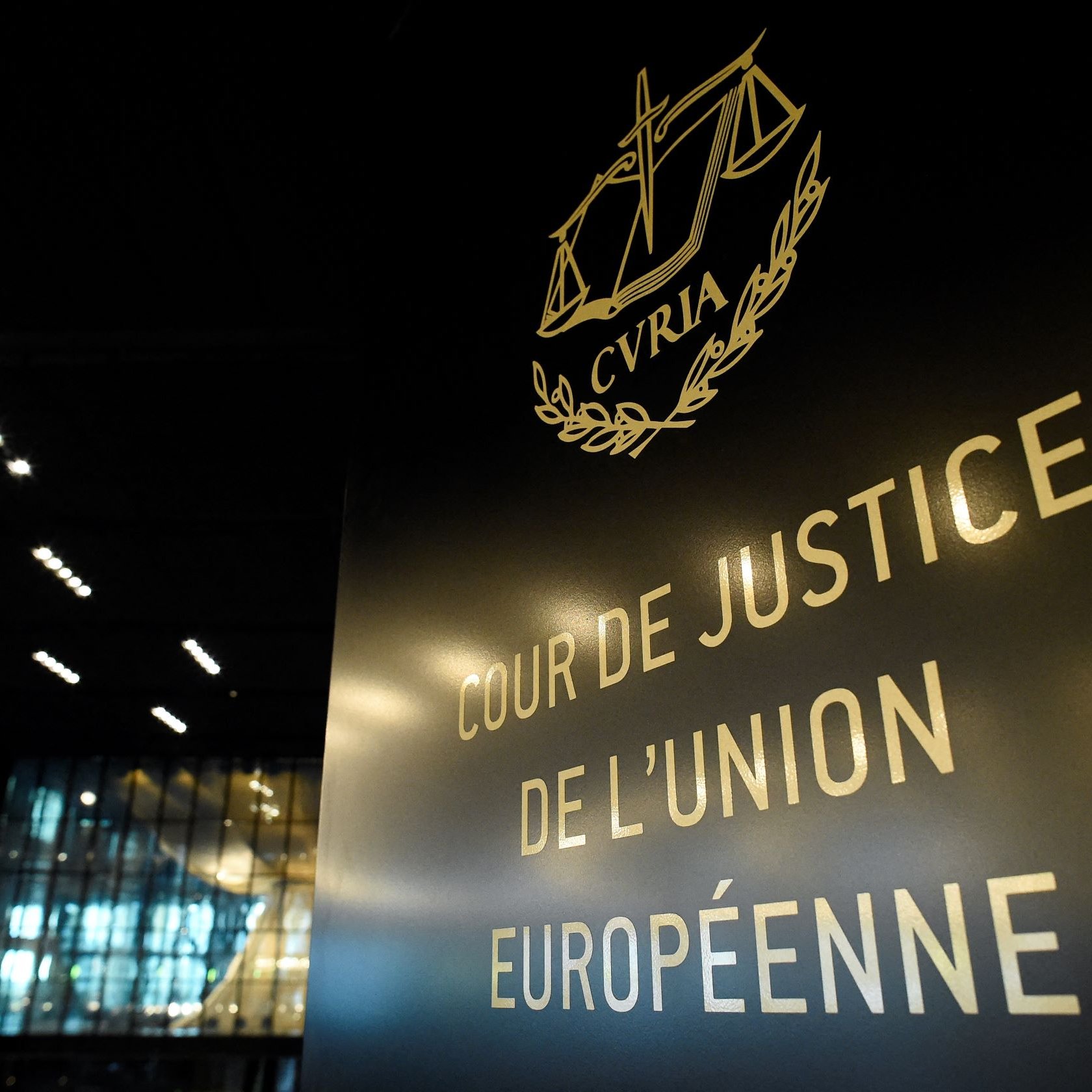 L'image montre un panneau indiquant "COUR DE JUSTICE DE L'UNION EUROPÉENNE". Le panneau est stylisé avec des éléments graphiques, notamment une représentation de la balance de la justice. En arrière-plan, on peut voir une architecture moderne et intérieure, probablement d'un bâtiment officiel. L'éclairage est subtil, mettant en valeur le texte et le design du panneau.