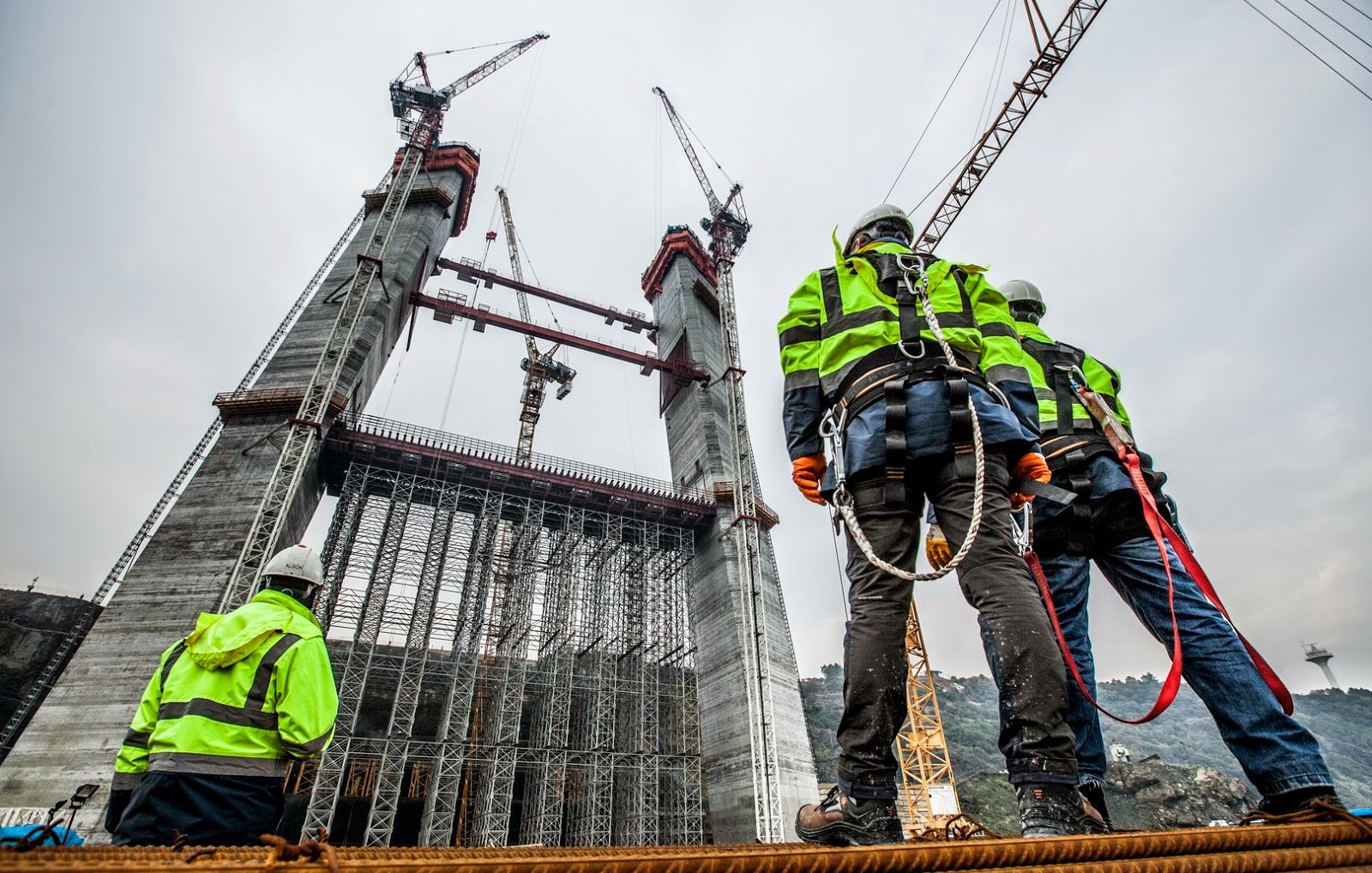 L'image montre une scène de construction avec plusieurs travailleurs vigilants observant une structure imposante en cours d'édification. Les ouvriers portent des vestes de sécurité à rayures et des casques, et sont accrochés avec des harnais de sécurité. En arrière-plan, on peut voir des grues et des éléments de construction en cours d'assemblage, tandis que le ciel est nuageux, suggérant une ambiance de travail intense sur le chantier.
