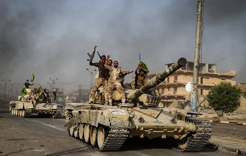 L'image montre des tanks avancant sur une route dans un environnement urbain. Plusieurs soldats sont visibles sur les tanks, certains levant les bras en signe de victoire ou de célébration. L'arrière-plan présente des bâtiments endommagés et une atmosphère chargée de fumée, ce qui suggère un contexte de conflit. Les couleurs sont principalement terreuses, avec des teintes de vert et de beige des tanks et des uniformes des soldats.