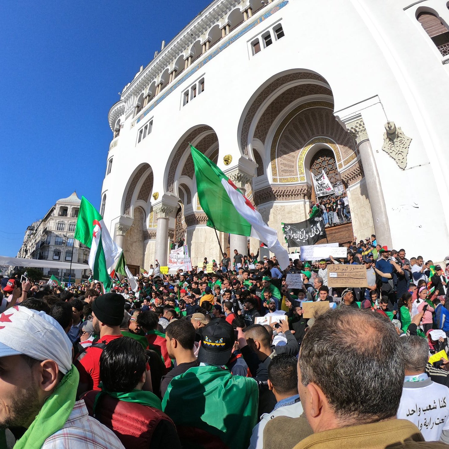 تظهر الصورة تجمعًا حاشدًا لمتظاهرين في مكان مفتوح، يحتشدون أمام مبنى تاريخي. يرفع المتظاهرون أعلام الجزائر بينما يحمل البعض لافتات تحمل شعارات. يتواجد عدد كبير من الأشخاص في الصورة، مما يبرز حماسهم ومشاركتهم الفاعلة. السماء زرقاء وظهور شعاع الشمس، مما يعكس أجواء نابضة بالحياة.