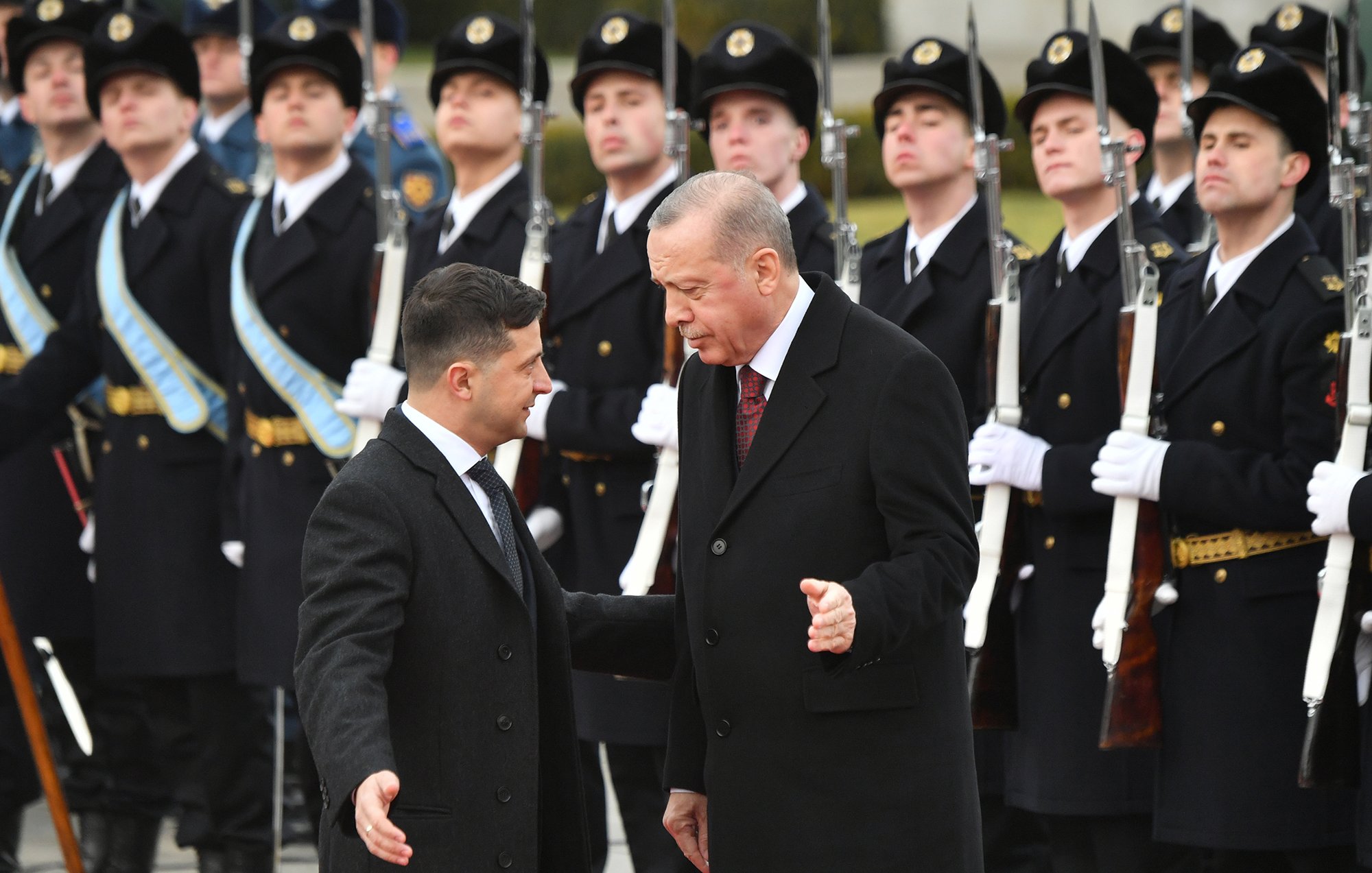 L'image montre deux hommes en discussion. L'un est vêtu d'un manteau noir, tandis que l'autre porte un costume avec une cravate. En arrière-plan, des soldats en uniforme présentent les honneurs, debout et alignés, avec des armes à la main. L'atmosphère semble solennelle, indiquant probablement une rencontre officielle ou un événement diplomatique.