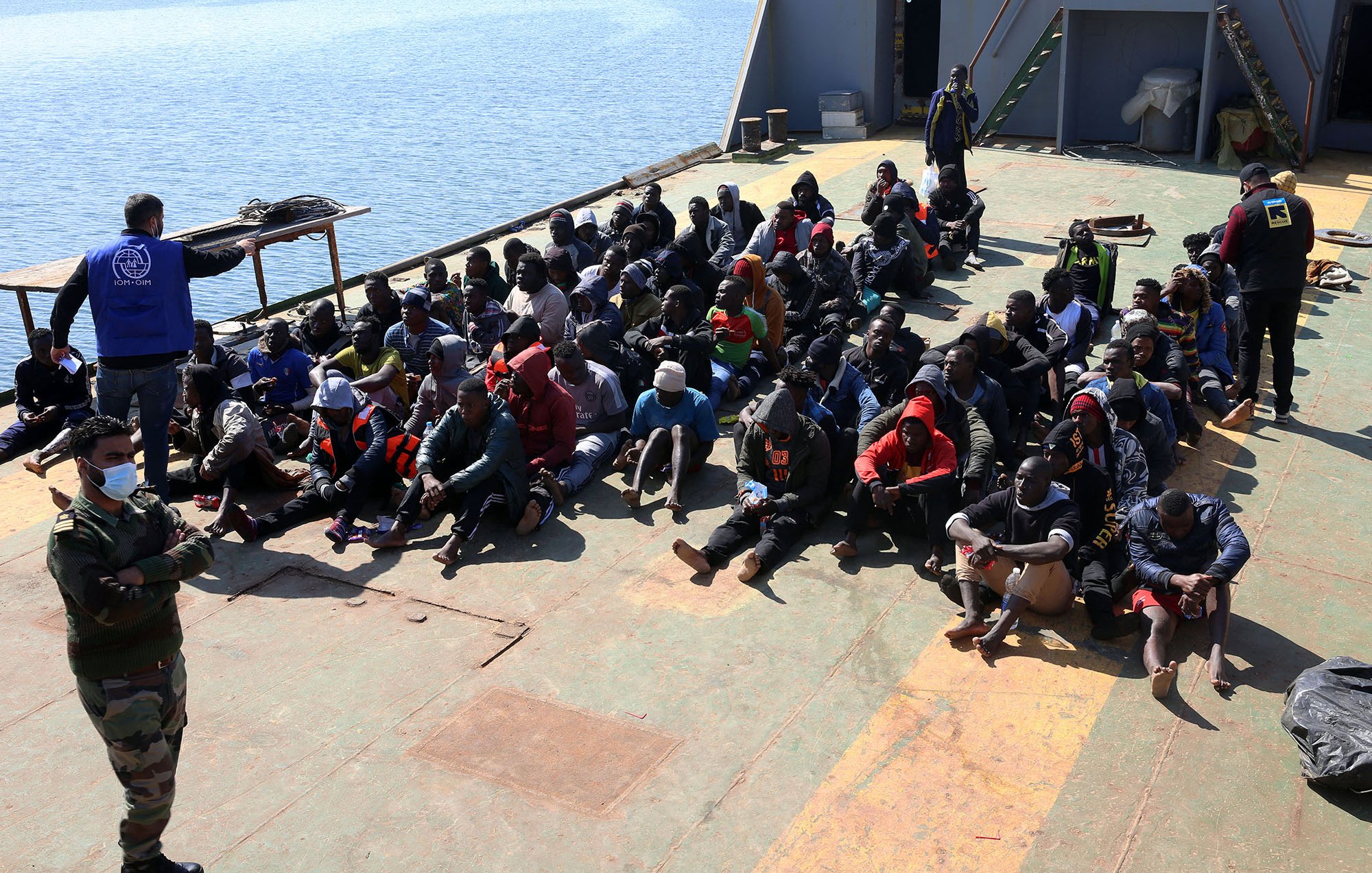 L'image montre un groupe de personnes assises sur le pont d'un bateau. Elles semblent être des migrants, plusieurs portant des vêtements chauds et des masques. À l'arrière, une personne en uniforme militaire observe la scène, tandis qu'un autre individu est vêtu d'un gilet portant un logo d'une organisation humanitaire. L'environnement semble être une zone portuaire, avec de l'eau visible à l'arrière-plan. L'atmosphère de l'image évoque une situation de contrôle ou d'assistance humanitaire.