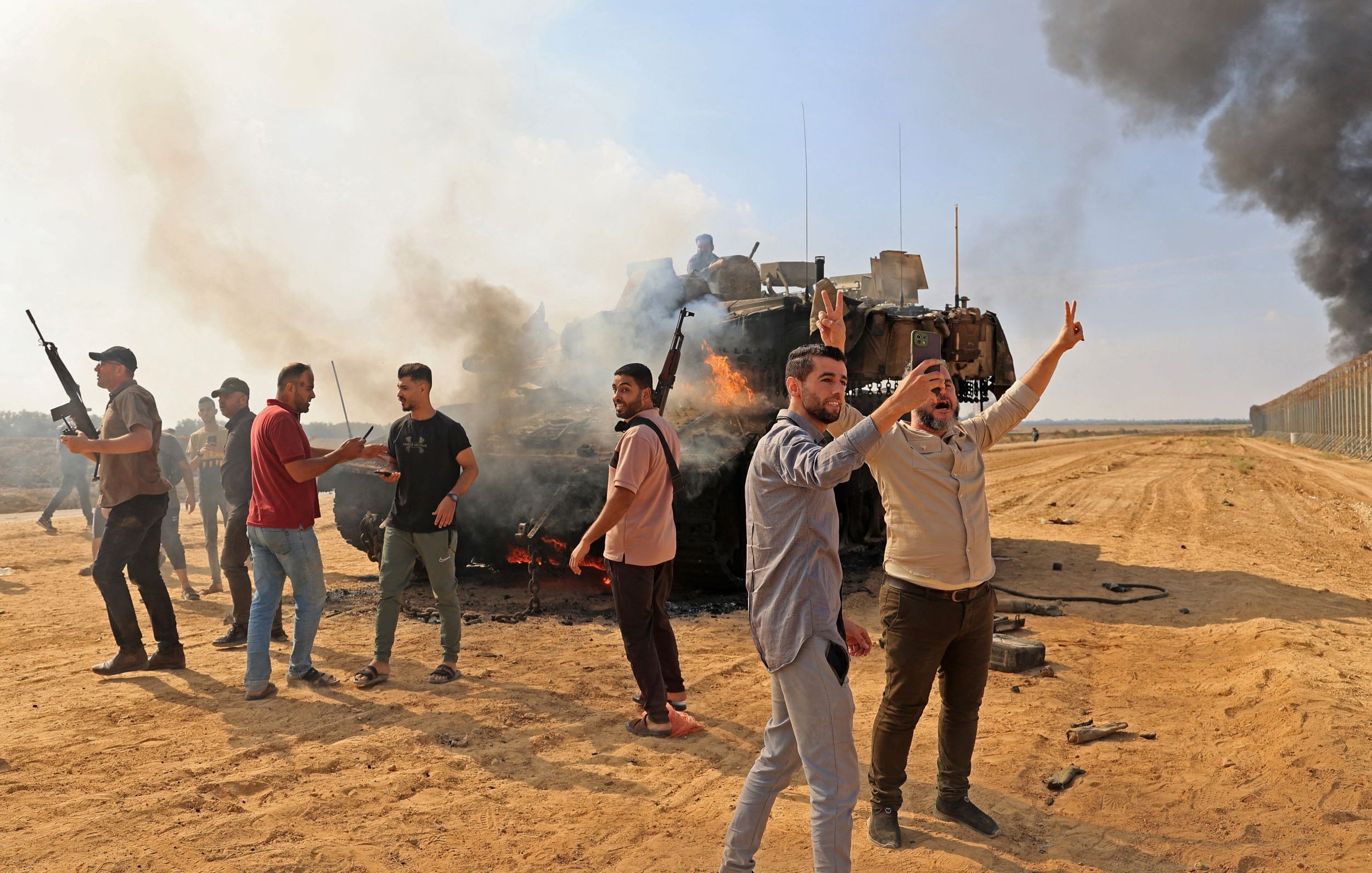 The image depicts a chaotic scene in a sandy area where a group of individuals appears to be celebrating or protesting near a burning military vehicle. Smoke rises from the vehicle, creating a dramatic backdrop. Some people are visible holding firearms, while others are raising their hands, possibly in a gesture of victory. The environment suggests a conflict or demonstration, with a tense atmosphere reinforced by the presence of smoke and the military vehicle. The landscape is open, with a barrier visible in the background, indicating a border or contested area.