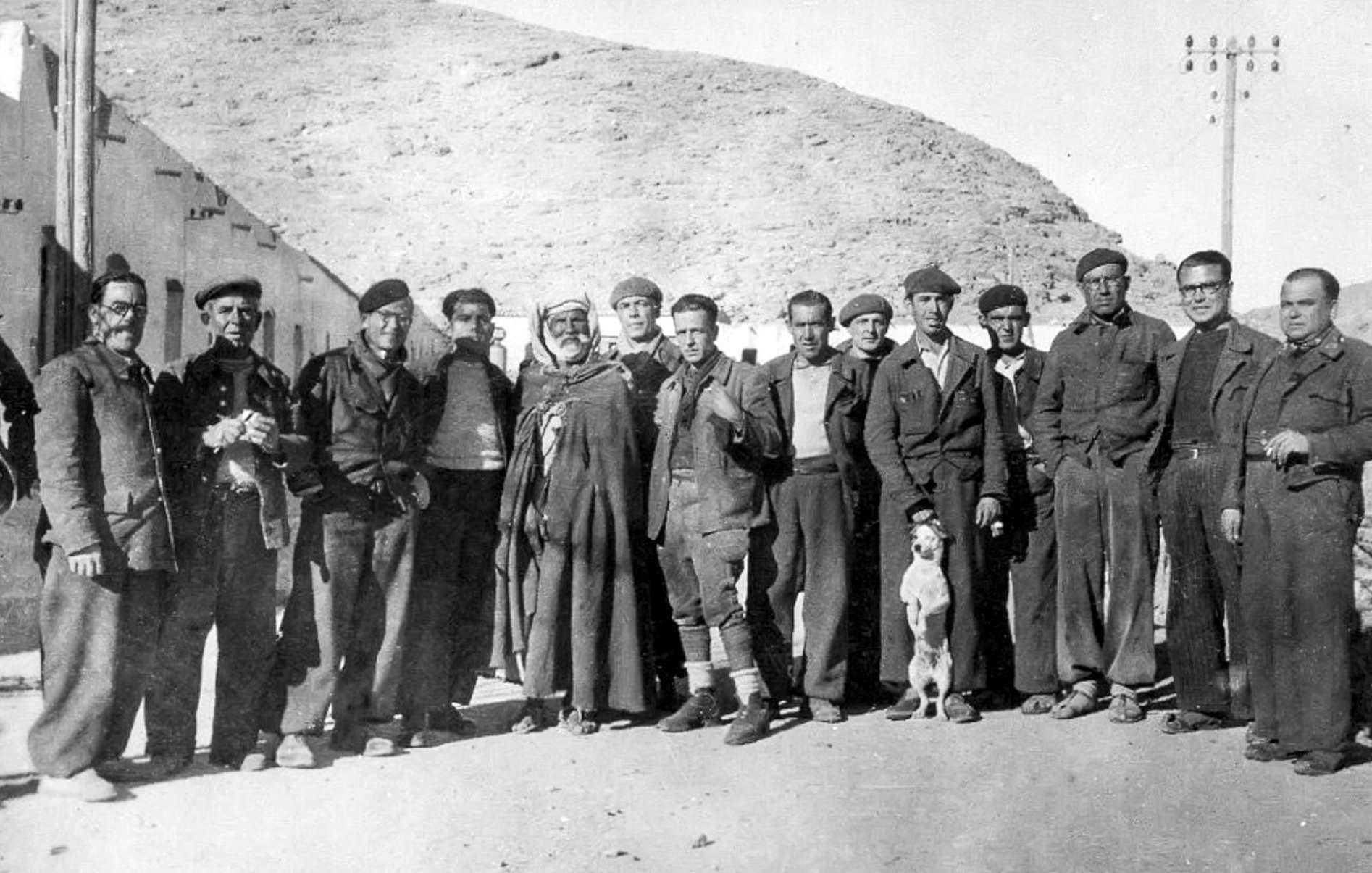 الصورة تظهر مجموعة من الأشخاص الذين يقفون معاً في مكان مفتوح، يبدو أنهم مجموعة تاريخية أو ربما فريق عمل. يرتدي معظمهم ملابس خاصة، مثل الزي العسكري أو ملابس العمل، وبعضهم يحمل أكواباً أو أدوات. في وسط المجموعة يوجد رجل يرتدي غطاء رأس تقليدي. هناك أيضاً كلب صغير يقف بين الأشخاص. تبدو الخلفية جبلية، مما يشير إلى أنهم في منطقة ريفية أو جبلية. تعكس الصورة أجواء من زمن بعيد وتحمل طابعاً تاريخياً.