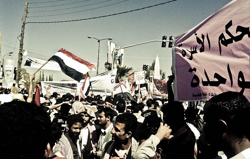 L'image montre une manifestation animée avec une grande foule de personnes. Des drapeaux flottent dans l'air, en particulier un drapeau qui semble être celui du Yémen. On peut voir des hommes et des femmes rassemblés avec des expressions déterminées. Des pancartes sont tenues par des manifestants, certaines comportant des inscriptions en arabe. L'atmosphère est dynamique, reflétant le désir de changement ou de revendication au sein de la foule. Les bâtiments environnants et les lignes électriques en arrière-plan ajoutent un contexte urbain à la scène.