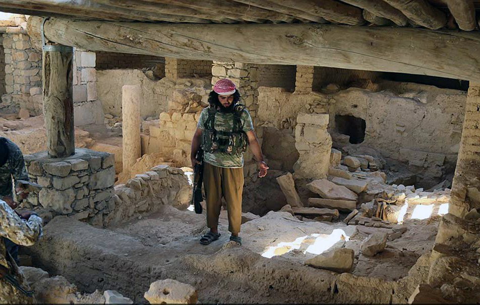 L'image montre un intérieur d'un bâtiment qui semble ancien et en ruine. On peut voir des murs en pierre et des débris éparpillés sur le sol. Un homme, habillé en vêtement militaire avec un bandeau, se tient au centre de la pièce, tandis que deux autres personnes, cachées en partie, l'observent. La lumière entre par des ouvertures, créant un contraste entre les ombres et les zones illuminées dans cet environnement désolé.