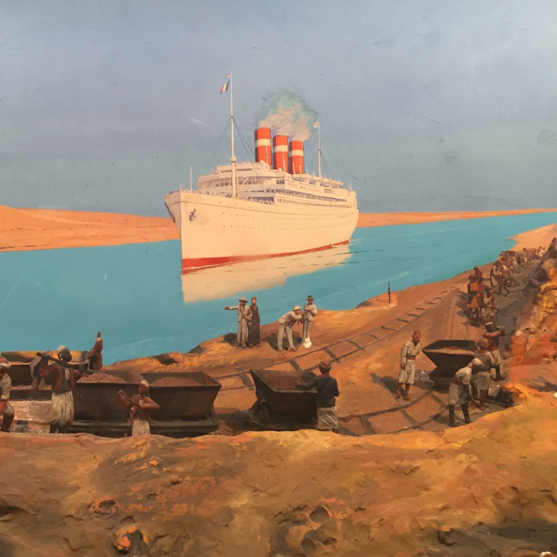 L'image représente une scène historique dans un environnement maritime. On voit un grand paquebot avec trois cheminées rouges amarré près d'une berge, probablement dans un canal ou un fleuve. Au premier plan, plusieurs hommes travaillent sur la rive, transportant des charges, probablement des matériaux ou des marchandises. À l'arrière-plan, le paysage désertique montre des dunes de sable sous un ciel bleu, créant un contraste avec l'eau turquoise. Cette image évoque l'activité maritime et le travail acharné des ouvriers dans un contexte industriel.