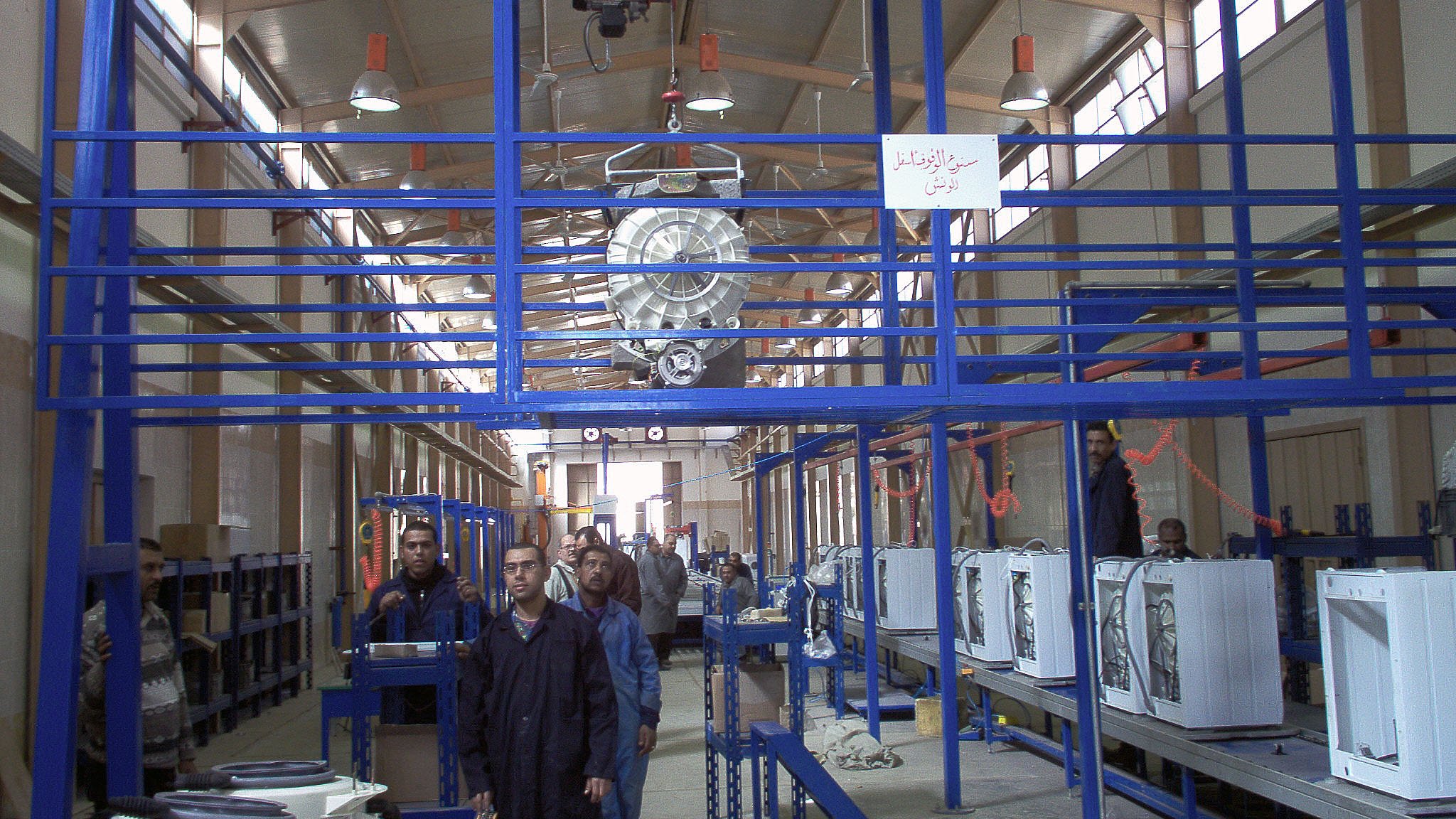 L'image montre un environnement d'atelier ou d'usine. On peut y voir des structures en métal, probablement des systèmes de rangement ou de support. Il y a des personnes travaillant dans cet espace, accompagnées d'équipements comme des appareils électroménagers, qui semblent être en phase de montage ou de préparation. L'éclairage est suffisant, et l'agencement des machines et des travailleurs suggère une ligne de production ou un processus d'assemblage. On note également des éléments rigides de la construction qui maintiennent l'espace organisé.