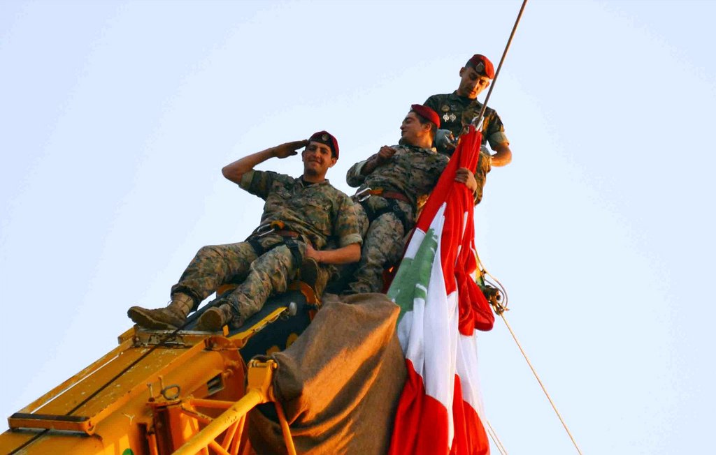 L'image montre trois soldats en uniforme, perchés en haut d'une grue. Ils sont en train de saluer tout en tenant un grand drapeau, probablement celui du Liban. Les soldats affichent un air déterminé et solennel, et l'ambiance semble festive, suggérant une célébration ou un événement important. Le ciel est clair, ce qui met en valeur la scène.