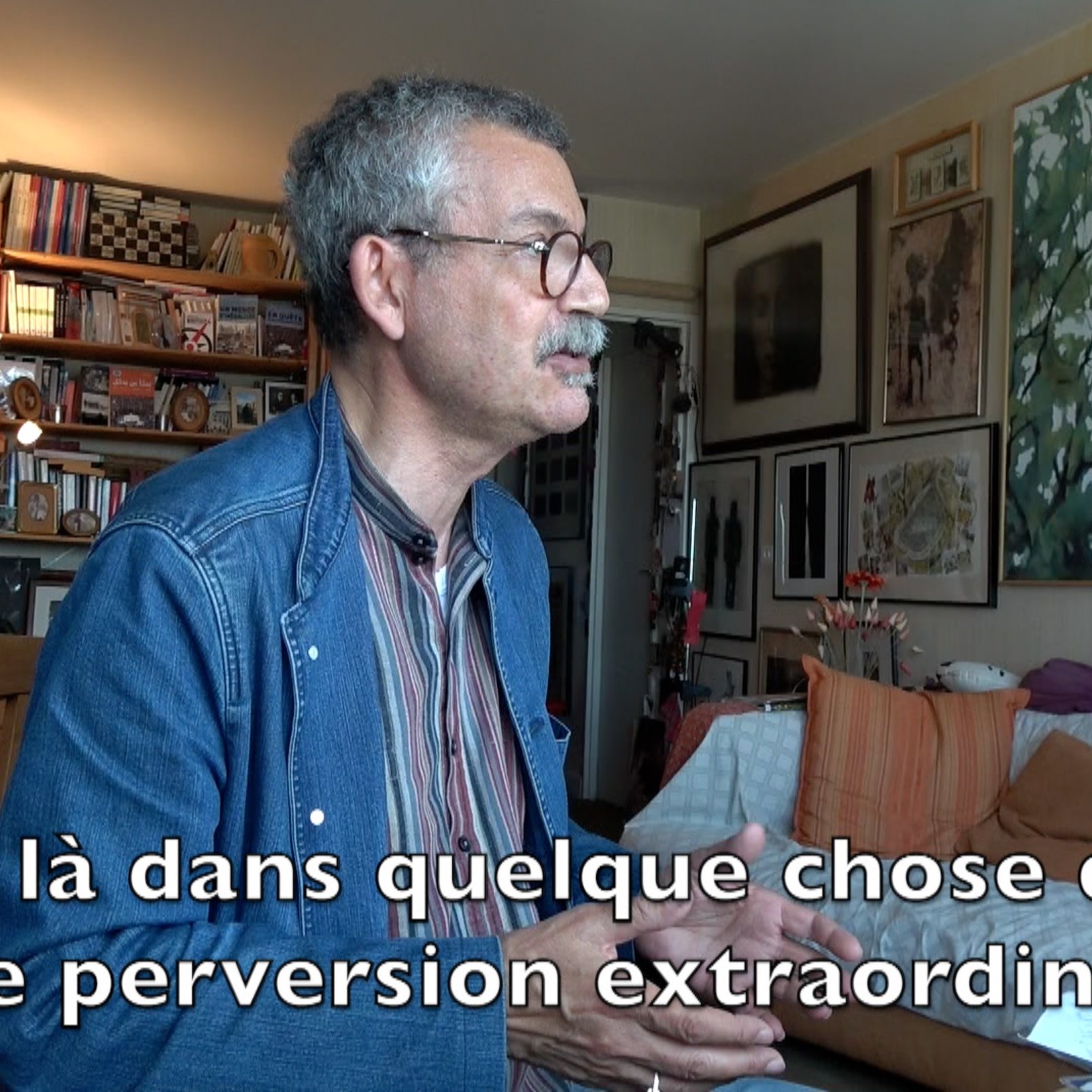 L'image montre un homme assis à une table, parlant avec animation. Il semble s'exprimer sur un sujet profond, probablement lié à des thèmes intellectuels ou sociaux. En arrière-plan, on peut voir des étagères remplies de livres et plusieurs œuvres d'art accrochées aux murs, ce qui indique un environnement artistique et studieux. Le texte sur l'image suggère qu'il aborde des concepts de manière critique, impliquant une réflexion sur des questions de moralité ou de société.
