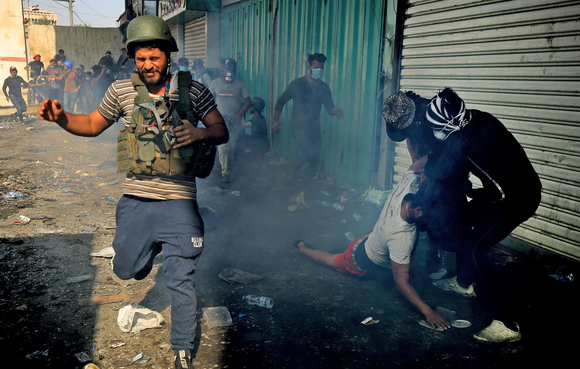 تصویر نشان‌دهنده یک صحنه پرتنش و شلوغ است. در foreground، یک مرد در حال دویدن به سمت دوربین است و ظاهراً در حال فرار از یک موقعیت خطرناک است. او کلاه‌خود و جلیقه نظامی به تن دارد. در پس‌زمینه، چند نفر دیگر دیده می‌شوند که در حال تعامل با یکدیگرند، یکی از آنها به زمین افتاده است. اطراف این افراد، دود و بخار به‌دلیل درگیری یا آشوب وجود دارد و زمین مملو از زباله و اشیای مختلف است. صحنه به وضوح فضای اضطراب و تنش را منتقل می‌کند.