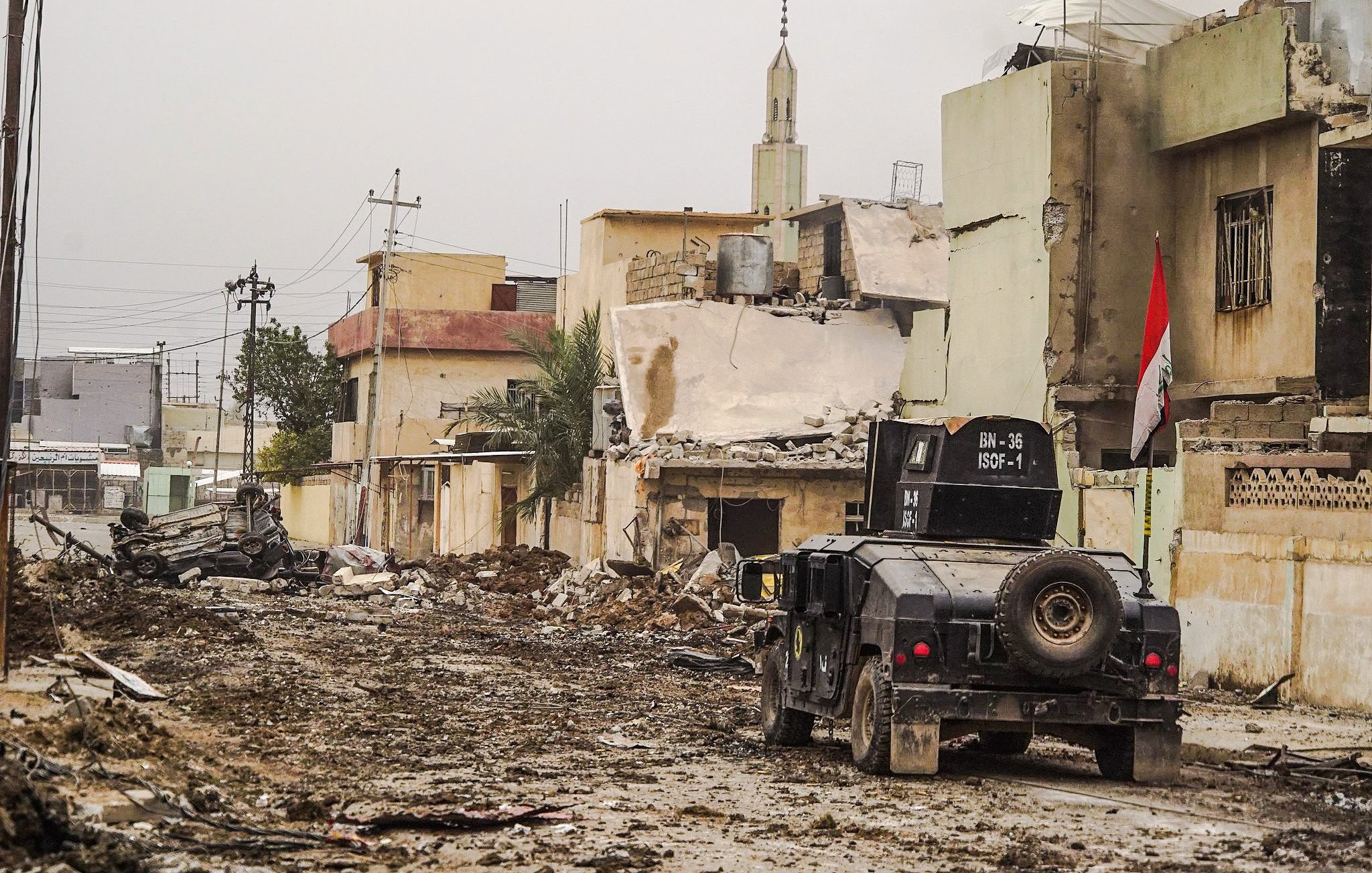 L'image montre une rue dévastée, probablement dans une zone de conflit. Au premier plan, on voit un véhicule militaire sur une route boueuse, entouré de débris. Les bâtiments en arrière-plan semblent endommagés, avec des murs effondrés et des structures incomplètes. Une mosquée est visible, avec son minaret se dessinant contre le ciel. Un drapeau irakien est aussi présent, indiquant que la scène se déroule en Irak. L'atmosphère générale semble sombre et dévastée, reflétant les conséquences d'un conflit armé.