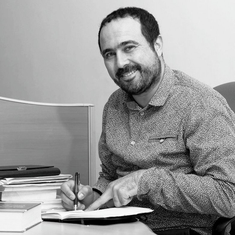 L'image montre un homme assis à un bureau, souriant et tenant un stylo dans une main tout en feuilletant un carnet ou un livre avec l'autre main. Des piles de livres sont présentes sur le bureau, ainsi qu'une tasse à café posée à côté de lui. L'environnement semble être un bureau, avec un fond minimaliste. L'image est en noir et blanc, ce qui lui donne un aspect classique.