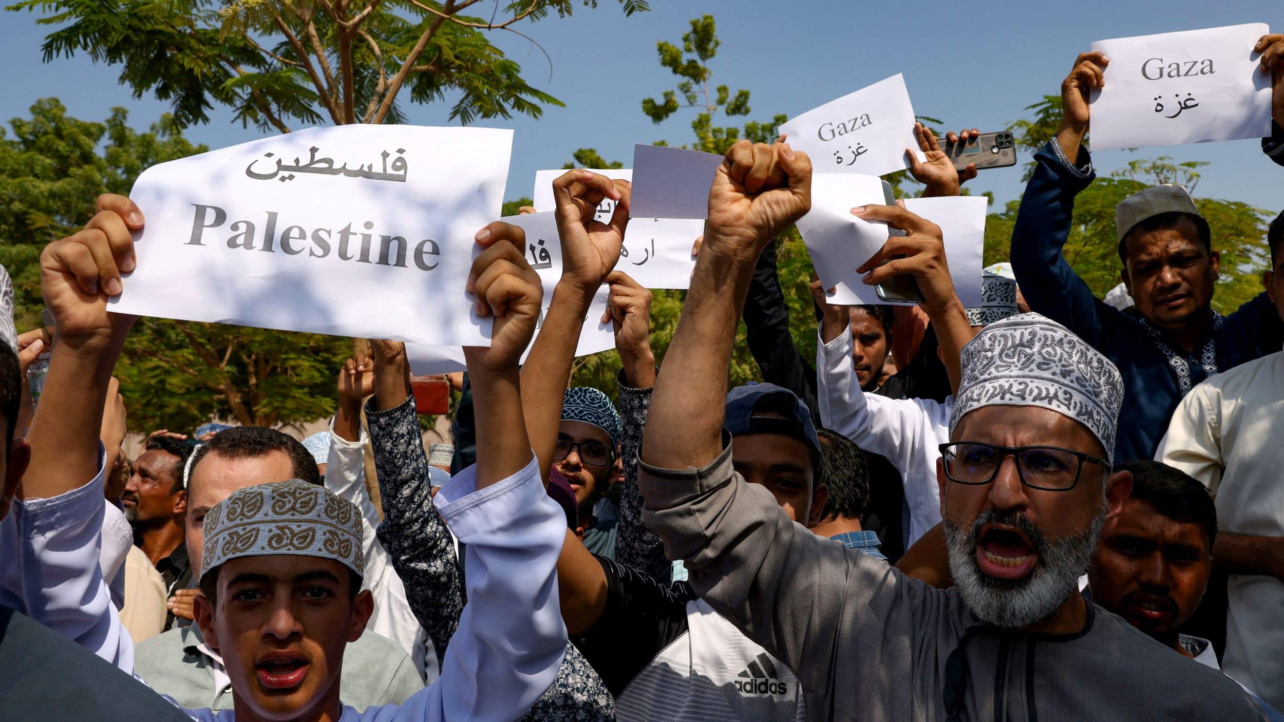 L'image montre un groupe de personnes tenant des pancartes en soutien à la Palestine et à Gaza. Les manifestants semblent rassembler pour exprimer leur solidarité, avec des expressions déterminées et des chants. Les pancartes affichent des mots en arabe ainsi qu'en anglais, reflétant un message de soutien à la cause palestinienne. L'environnement semble être en plein air, probablement lors d'une manifestation ou d'un rassemblement.