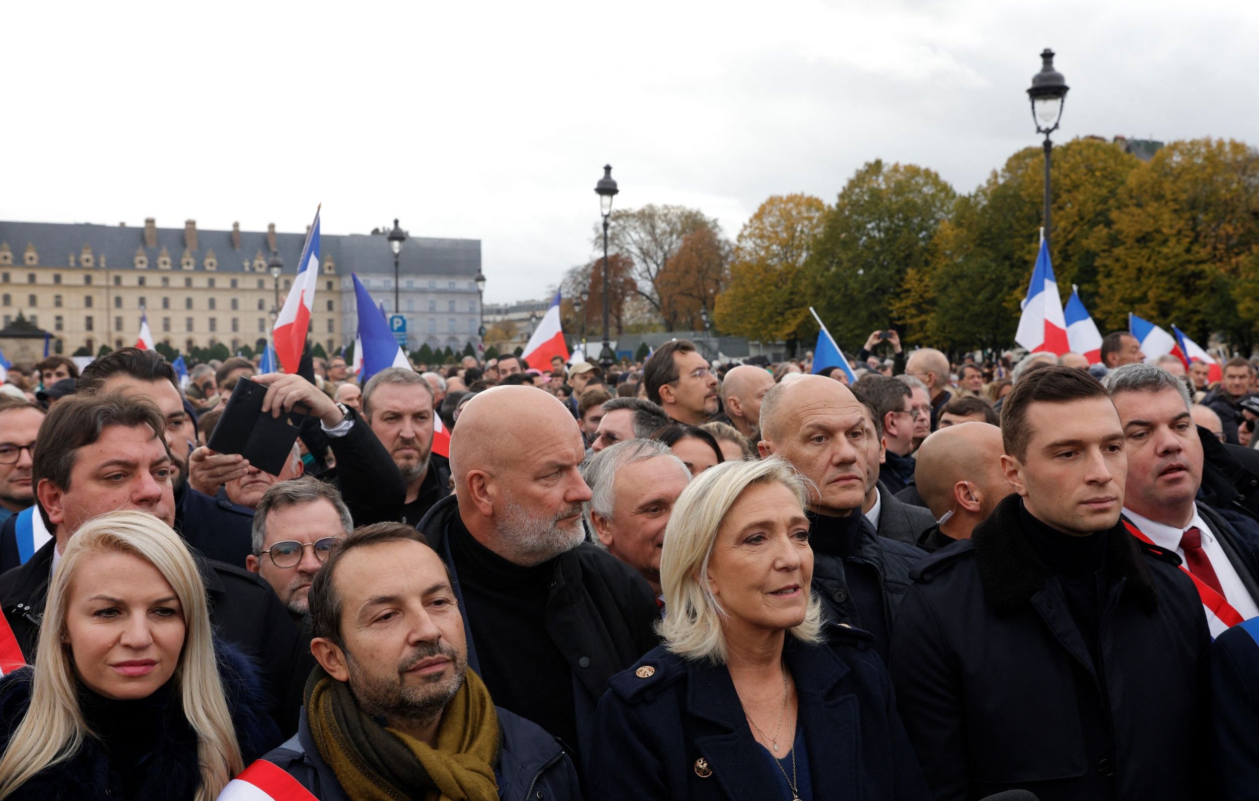 The image depicts a crowd of people gathered in an outdoor setting, likely at a political event or rally. Many individuals are holding French flags, and the expressions on their faces suggest a mix of determination and focus. Prominent figures can be seen at the forefront, wearing formal attire. The background features historic architecture and trees, indicating an urban environment. The atmosphere appears charged, reflecting the significance of the gathering.
