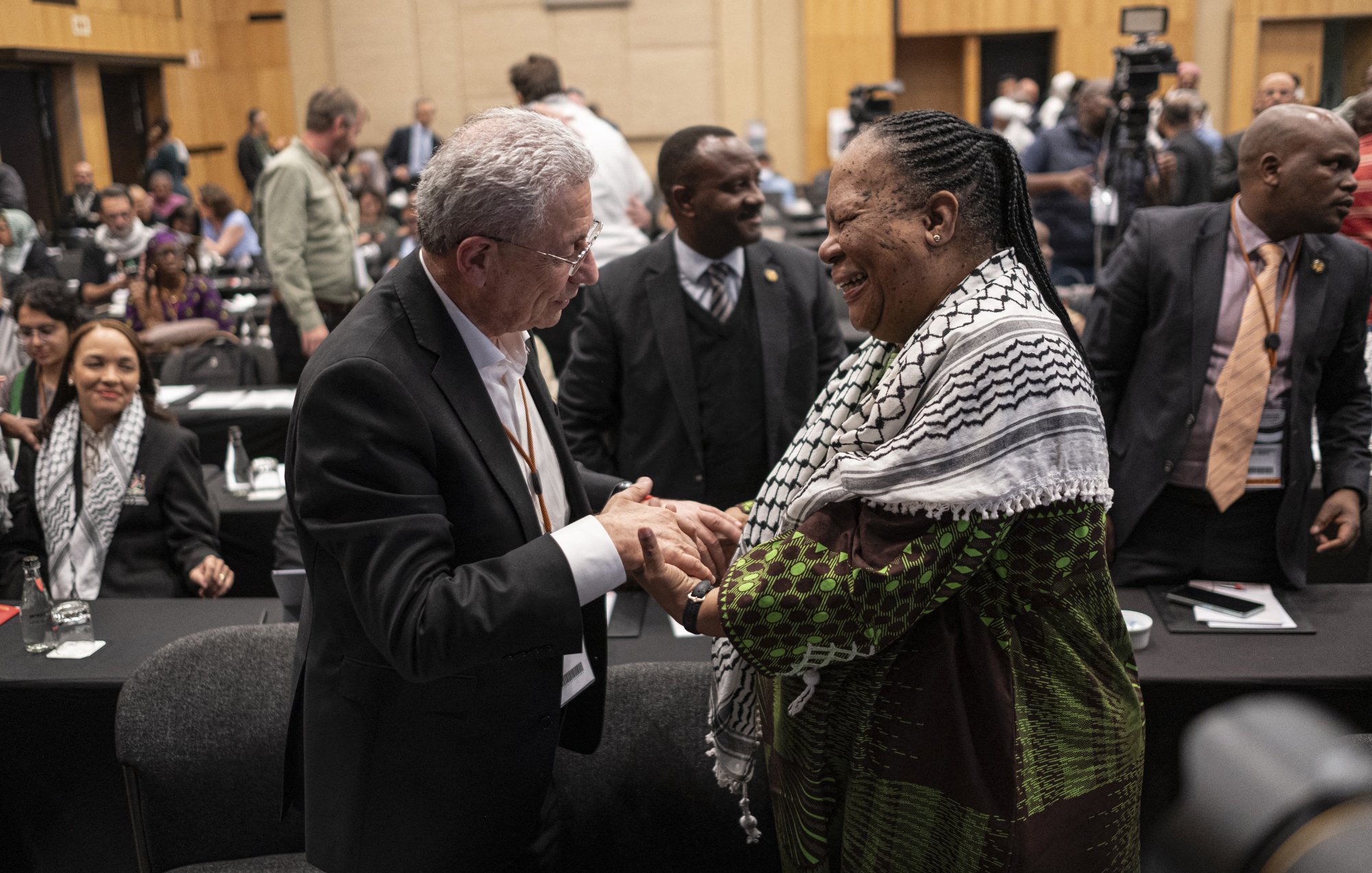 The image depicts a moment of interaction between two individuals at a conference or event. Both people are smiling and engaged in a handshake, suggesting a friendly and positive exchange. The person on the left is wearing a suit, while the individual on the right is dressed in traditional or cultural attire with a colorful shawl. In the background, there are other attendees, some seated and others standing, indicating a busy atmosphere typical of a conference. The setting appears to be indoors, likely within a large hall or meeting room.