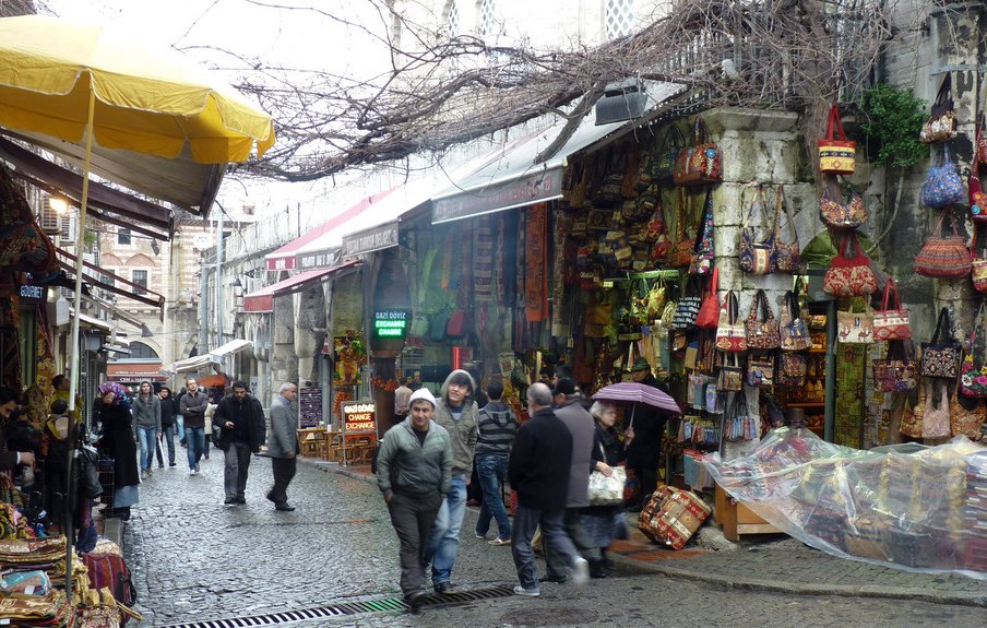 الصورة تُظهر زقاقاً ضيقاً في سوق شعبي، حيث تتعانق جوانب الطريق مع المحلات التي تبيع مجموعة متنوعة من السلع. تُظهر المحلات أرففاً مليئة بالأكسسوارات والحقائب الملونة، وقد تكون هناك بعض الزبائن يتجولون بين المتاجر. السماء ملبدة بالغيوم، وبعض الأشخاص يحملون مظلات، مما يشير إلى احتمال وجود مطر. يوجد أيضاً ظلال وزوايا تعكس أجواء الأسواق التقليدية.