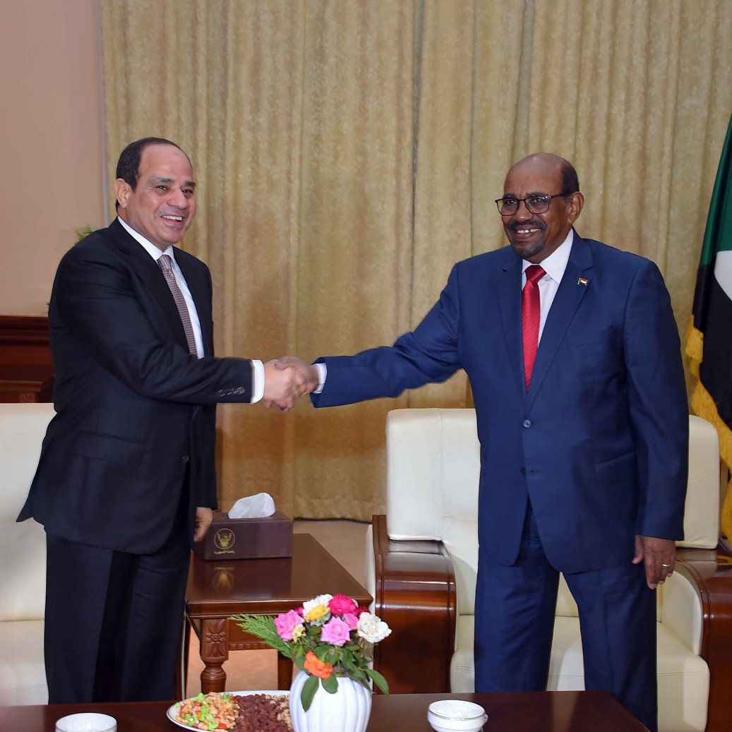 L'image montre deux hommes en train de se serrer la main dans un cadre de rencontre officielle. L'un porte un costume sombre et l'autre un costume bleu. Ils sont entourés d'un décor formel, comprenant des drapeaux et des éléments de décoration. La scène suggère un moment de dialogue ou de coopération entre les deux personnes, probablement des responsables politiques. Un vase avec des fleurs est posé sur la table entre eux.