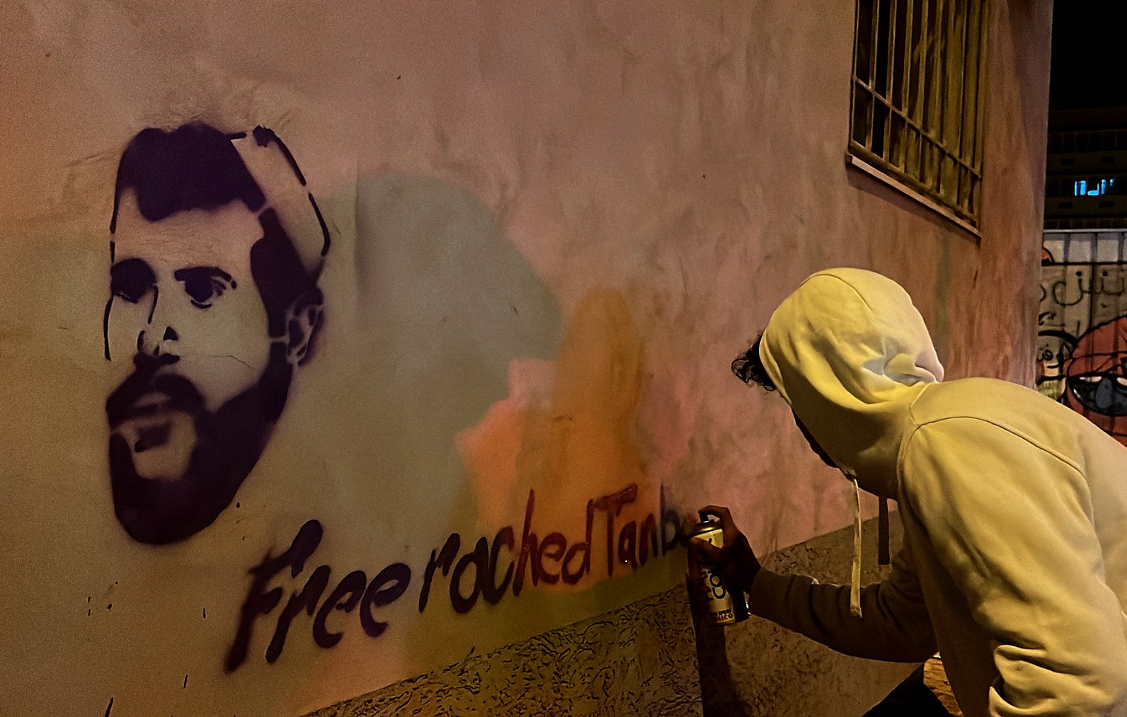 L'image montre une personne de dos, habillée d'un sweat à capuche blanc, en train de peindre un graffiti sur un mur. Le graffiti représente le visage d'un homme, probablement une figure connue, avec la phrase "Free roched Tank" écrite en dessous. La scène se déroule la nuit, et l'éclairage crée des reflets colorés sur le mur. On peut également apercevoir un portail ou une autre surface à proximité avec un autre graffiti en arrière-plan.