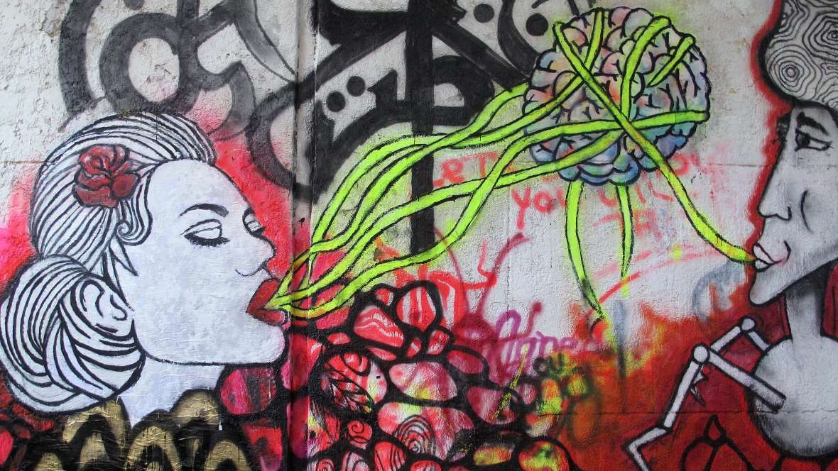L'image montre une œuvre de street art colorée sur un mur. À gauche, il y a le portrait d'une femme avec un rose dans les cheveux, qui semble "parler" ou "chanter". À droite, un homme est représenté, affichant une expression sérieuse. Entre les deux, des lignes vertes émanent de la tête de la femme, semblant évoquer une connexion ou un échange de pensées, illustrées par un cerveau stylisé en arrière-plan. L'ensemble est dominé par des couleurs vives et des motifs abstraits, créant une atmosphère vibrante et dynamique.