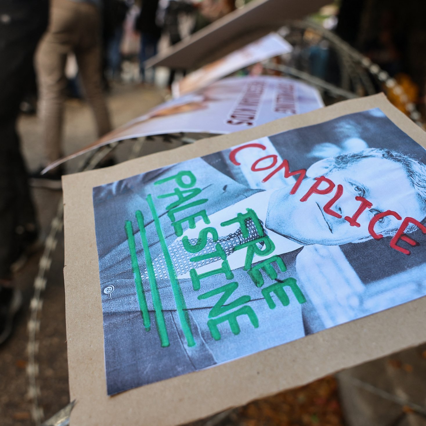 L'image montre des affiches placées sur une barrière, probablement dans le cadre d'une manifestation. L'une des affiches présente un portrait en noir et blanc avec les mots "COMPLICE" et "FREE PALESTINE" écrits en rouge et vert. L'environnement semble être celui d'une manifestation ou d'un rassemblement, avec plusieurs personnes en arrière-plan.