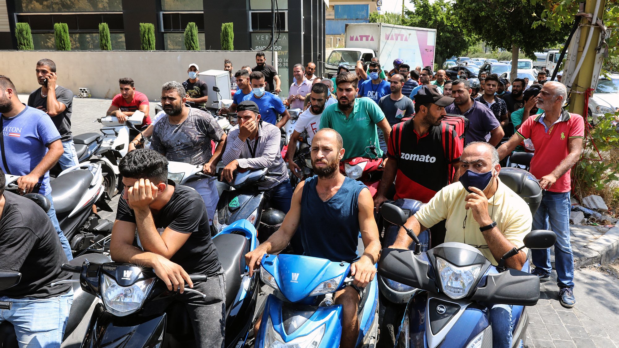 تظهر الصورة مجموعة من الأشخاص يتجمعون أمام مركز ما، حيث يبدو أنهم في انتظار الحصول على شيء ما. معظمهم يركبون دراجات نارية، ويبدو عليهم التوتر والقلق. بعضهم يظهر علامات الإحباط، بينما آخرون يتحدثون مع بعضهم. الخلفية تحتوي على مباني وأشجار، مما يشير إلى أن المشهد في منطقة حضرية.