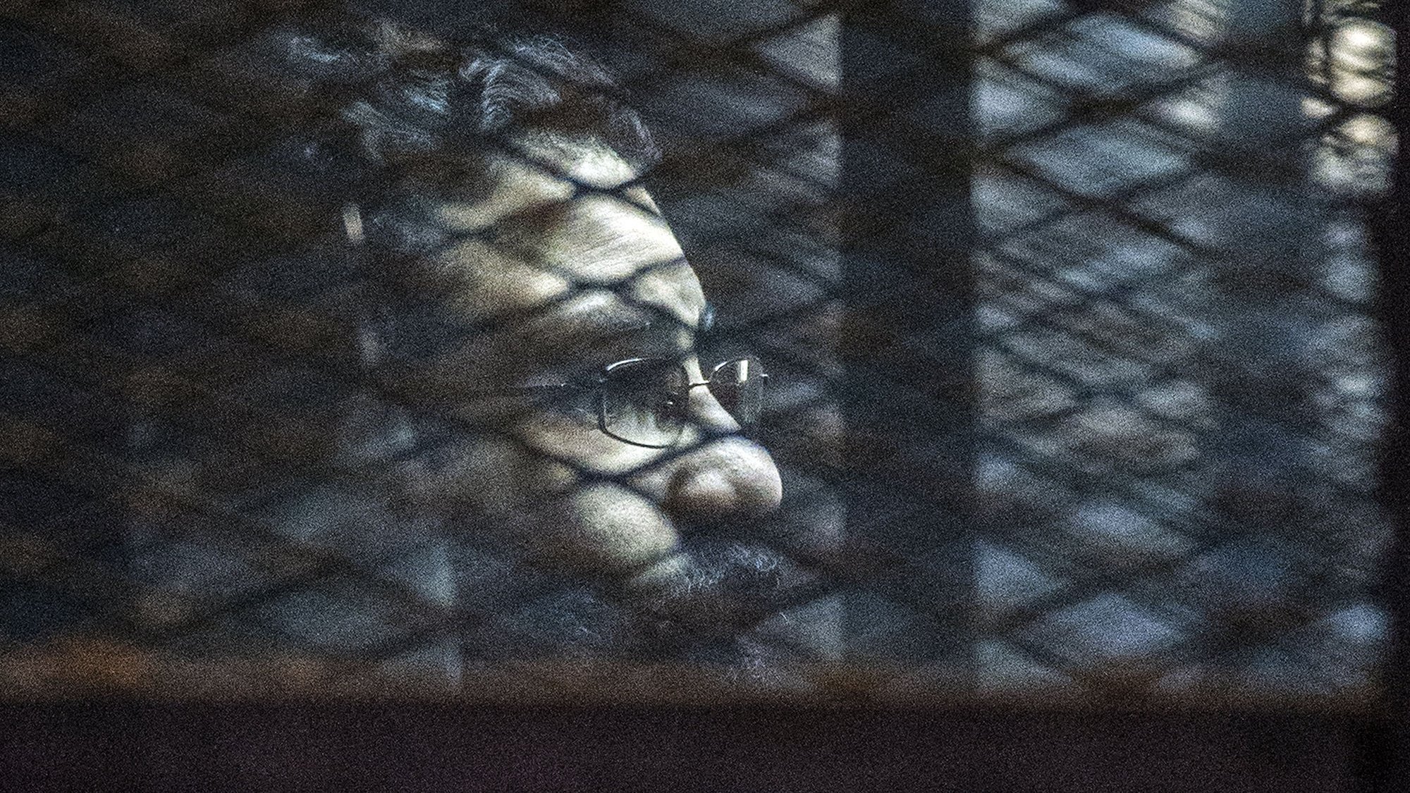 L'image montre un homme avec des lunettes et une barbe, dont le visage est partiellement obscurci par des ombres. On aperçoit des mailles ou des barres qui créent un effet de grille à l'avant, suggérant qu'il se trouve derrière une sorte de barrière ou de grillage. L'éclairage de la scène est sombre, contribuant à une ambiance mystérieuse.
