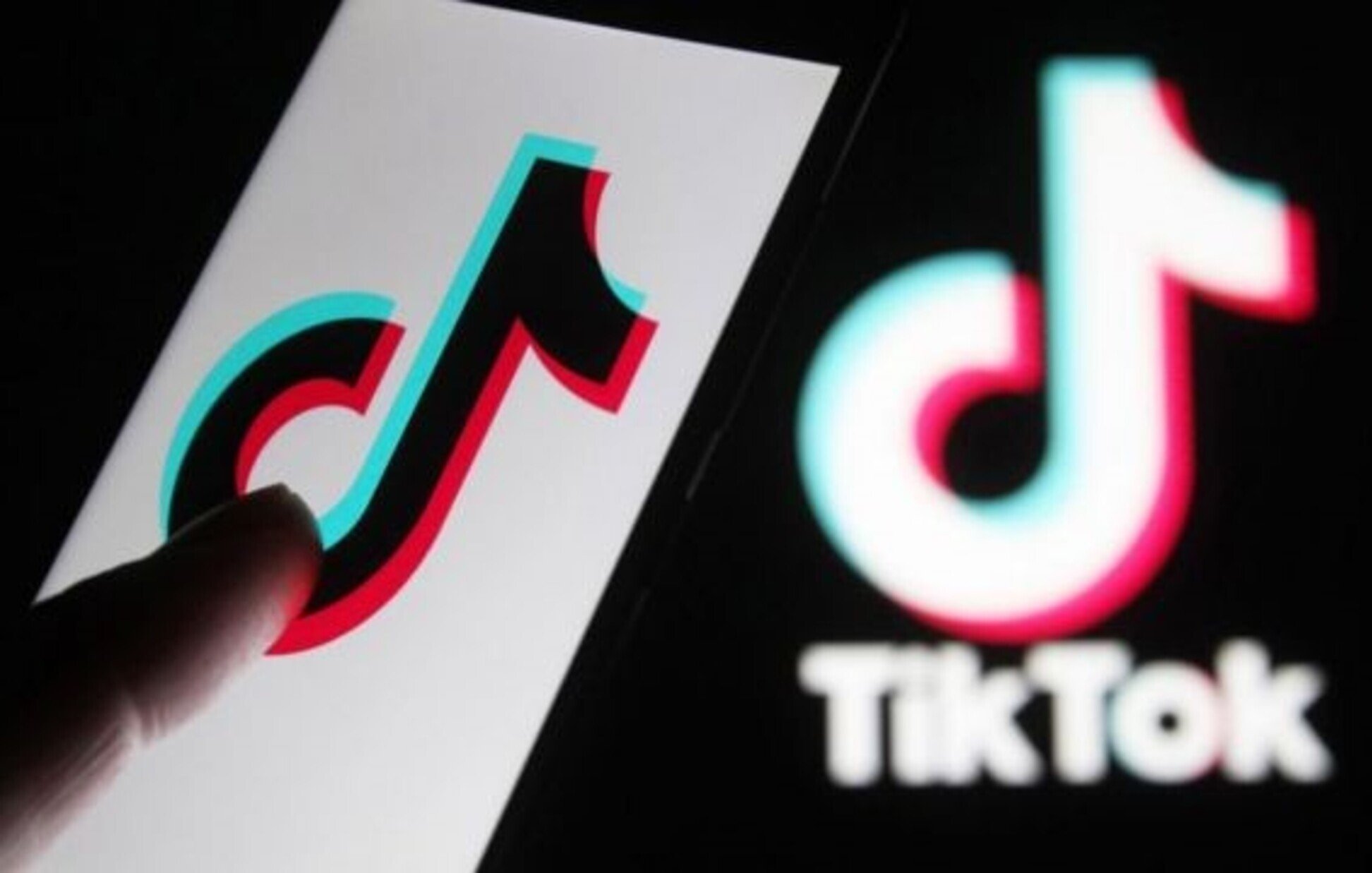 این تصویر یک گوشی هوشمند را نشان می‌دهد که لوگوی اپلیکیشن TikTok روی صفحه آن قابل مشاهده است. انگشت یک دست به سمت لوگو اشاره می‌کند. در پس‌زمینه، لوگوی TikTok به صورت بزرگ‌تر و با رنگ‌های آبی و قرمز دیده می‌شود. طراحی این لوگو به‌صورت مدرن و جذاب است و نشان‌دهنده‌ی ماهیت سرگرم‌کننده اپلیکیشن می‌باشد.