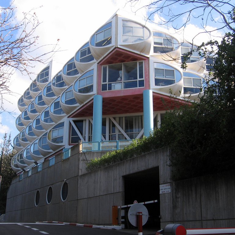 L'image montre un bâtiment moderne avec une architecture unique. Il se compose de plusieurs niveaux, avec une façade caractérisée par des fenêtres en forme de bulle ou de dôme. Les couleurs sont vives, notamment avec des éléments en rouge et en bleu. À l'avant, on peut voir un accès avec un panneau de signalisation et des éléments paysagers comme des arbres sur les côtés. Le bâtiment semble être intégré dans son environnement naturel, avec un design qui attire l'attention.