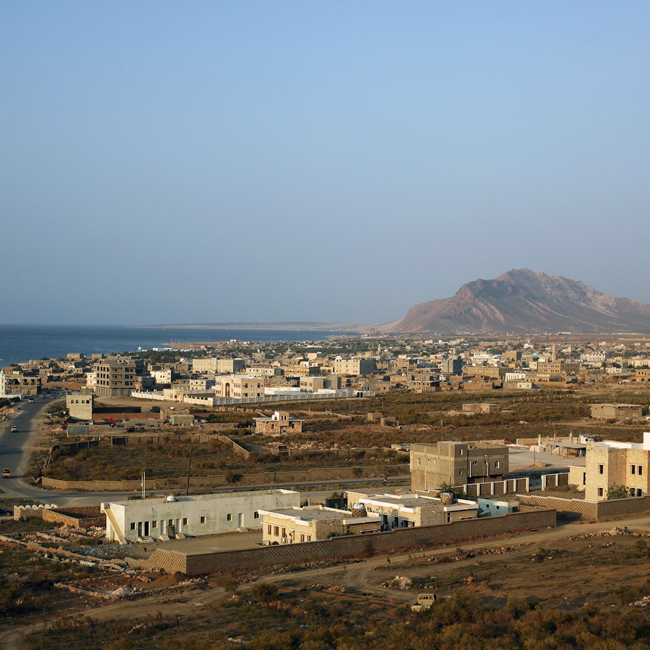 L'image montre un paysage côtier avec une vue sur une petite ville située près de la mer. On peut apercevoir des bâtiments de différentes tailles, certains modernes et d'autres plus traditionnels, dispersés sur une étendue de terrain. À l'arrière-plan, on distingue une montagne dont les contours se dessinent sous un ciel clair. La route serpente à travers la ville, menant à la mer. L'ensemble dégage une atmosphère paisible et semi-aride, typique des régions côtières.