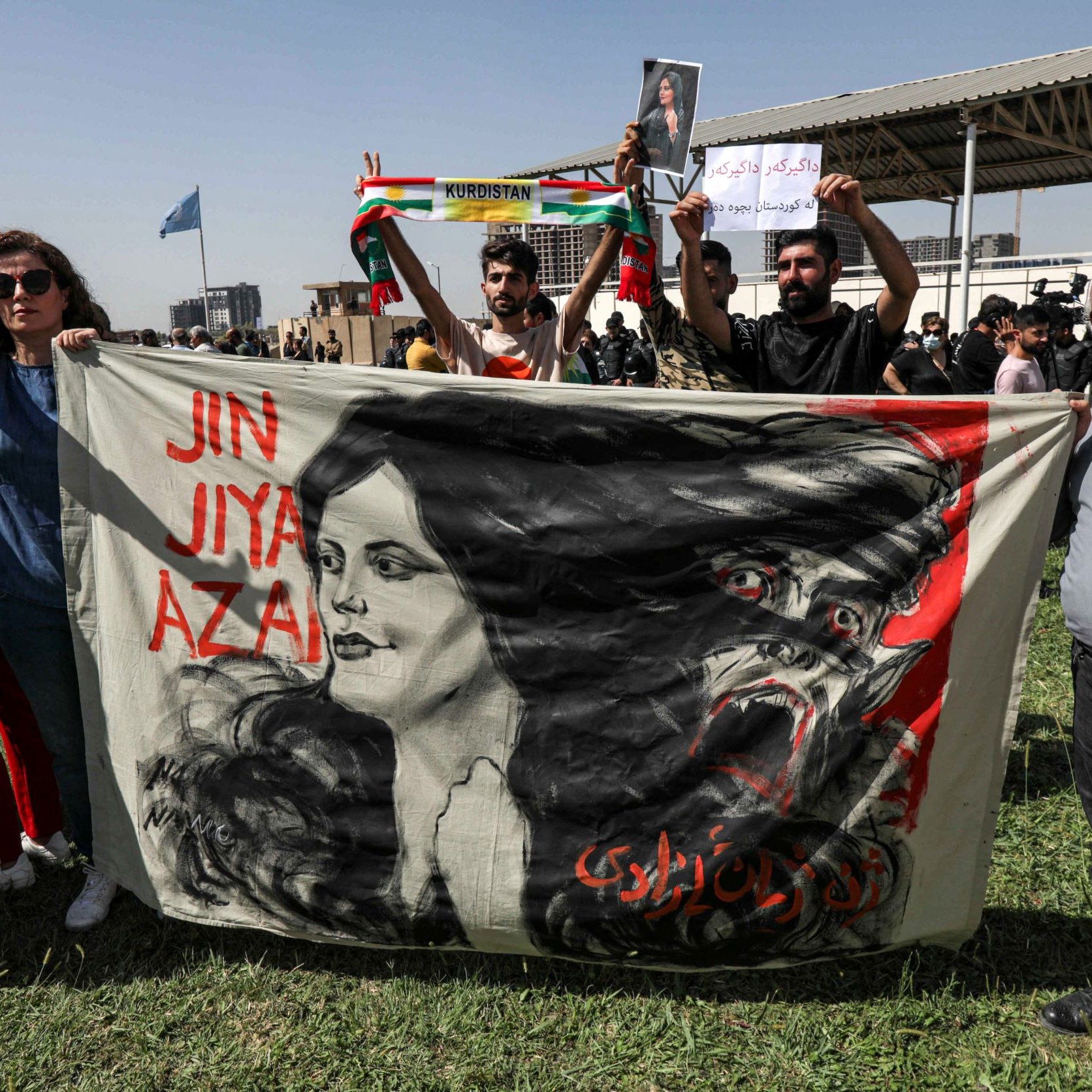 L'image montre une scène de manifestation. Des personnes tiennent une grande bannière sur laquelle est représentée le visage d'une femme entouré de motifs dramatiques. Les manifestants affichent des signes de paix avec leurs mains. On peut voir des drapeaux, probablement kurdes, en arrière-plan. L'atmosphère semble chargée d'émotion et de détermination, suggérant un rassemblement pour une cause importante.