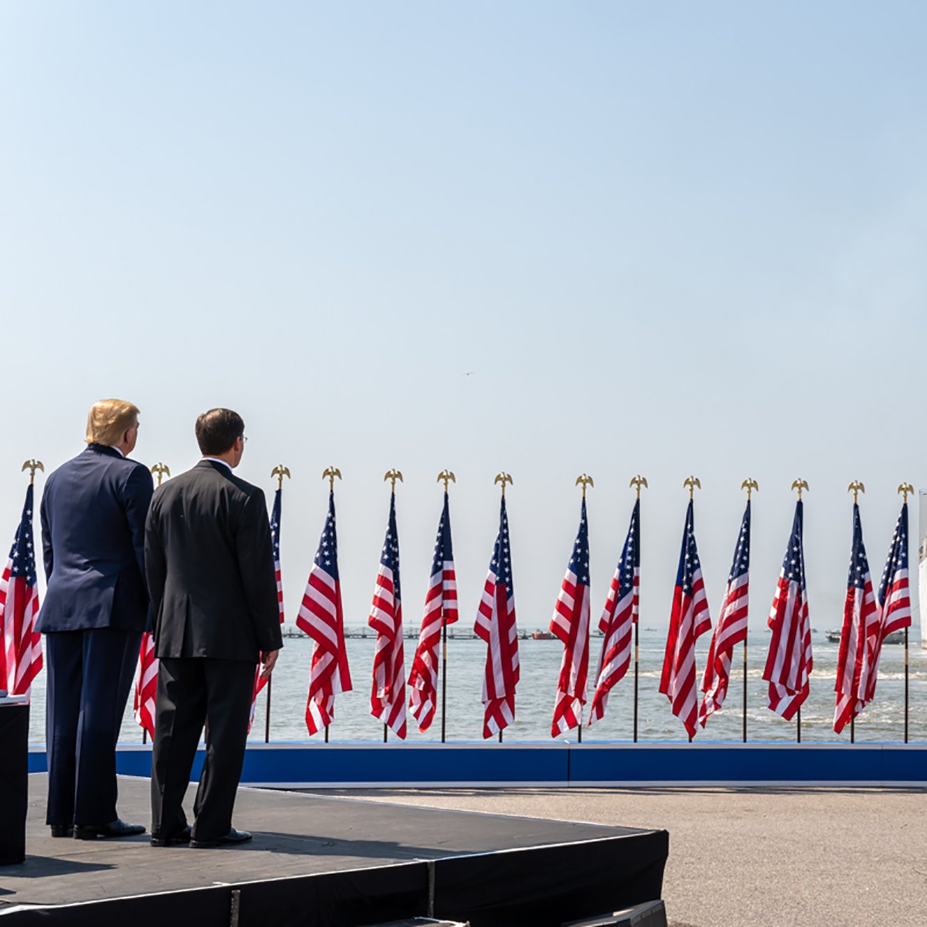 L'image montre une scène avec deux hommes de dos, face à un grand navire hospitalier qui arrive. Le navire est identifiable grâce à sa croix rouge sur le côté. Au premier plan, un podium est visible, entouré de multiples drapeaux américains flottant au vent. Le ciel est clair et lumineux, suggérant une journée ensoleillée. L'atmosphère semble solennelle, probablement en lien avec un événement officiel ou une cérémonie.