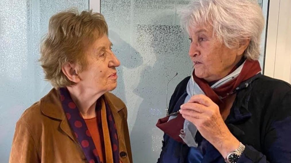 L'image montre deux femmes âgées engagées dans une conversation. Elles semblent amicales et souriantes. L'une porte une veste marron avec un foulard à pois, tandis que l'autre est habillée de manière similaire, avec une écharpe. L'arrière-plan est flou, mettant en valeur leur interaction.