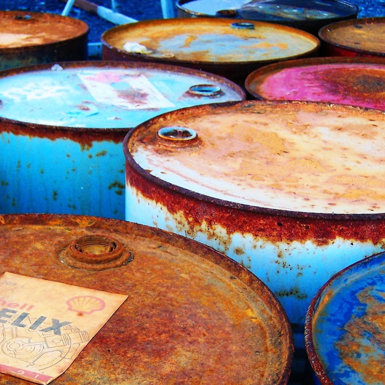 L'image montre plusieurs fûts en métal, probablement des fûts de pétrole ou d'huile. Ils sont empilés les uns à côté des autres, avec des couvercles rouillés et des étiquettes décolorées. La surface des fûts présente des signes d'oxydation, ce qui leur donne un aspect usé et ancien. Les couleurs variées du métal, allant du rouille au bleu clair, ajoutent à l'esthétique générale de la scène.