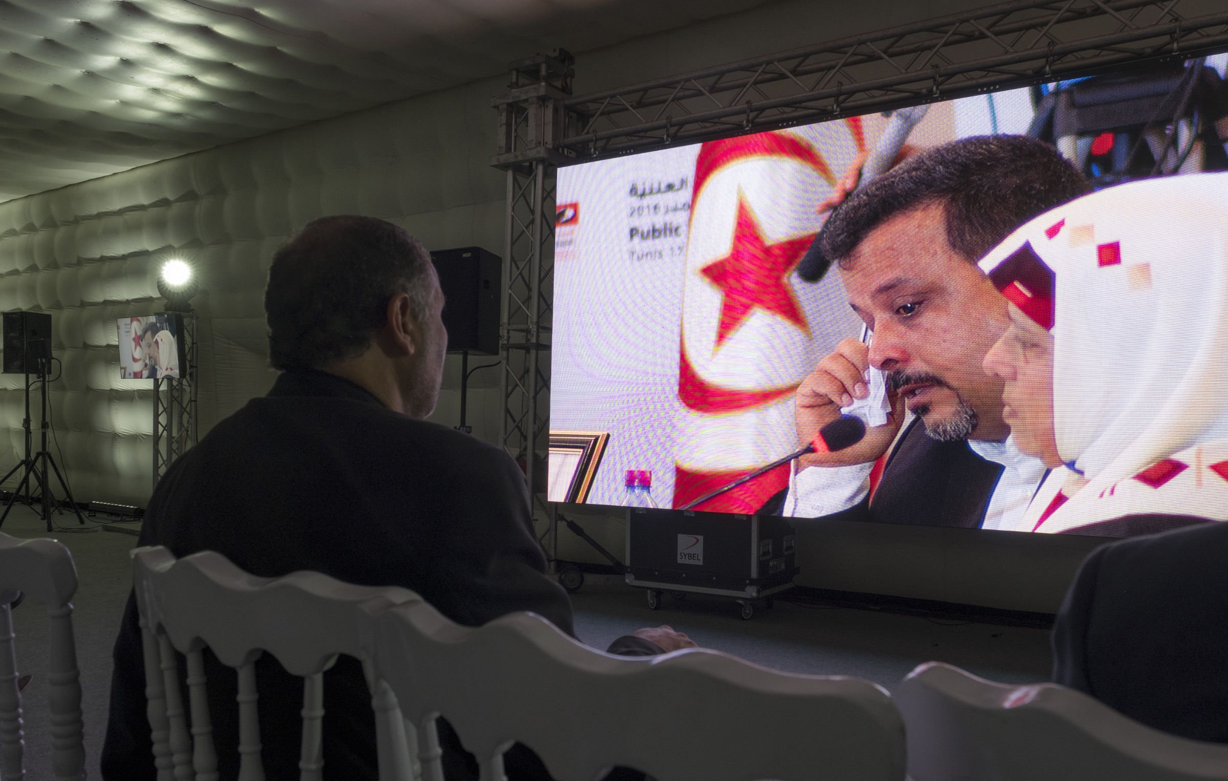 L'image montre un homme assis de dos, regardant un grand écran. Sur l'écran, on peut voir un homme parlant, avec une femme à ses côtés. L'ambiance semble formelle, peut-être liée à un événement officiel ou une conférence. L'éclairage et le décor sont modernes, avec des panneaux lumineux et des équipements audiovisuels visibles en arrière-plan. La scène dégage une atmosphère de concentration et d'attention.