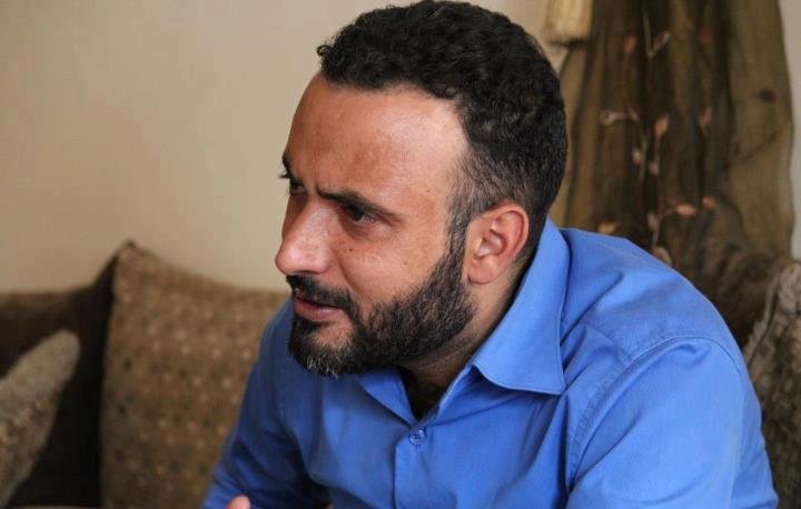 L'image montre un homme assis, avec une chevelure bouclée et une barbe bien taillée. Il porte une chemise bleu vif et semble en train de discuter ou de réfléchir, avec une expression sérieuse. L'arrière-plan est flou, mais on peut voir un canapé et quelques coussins. L'ambiance semble informelle et conviviale.