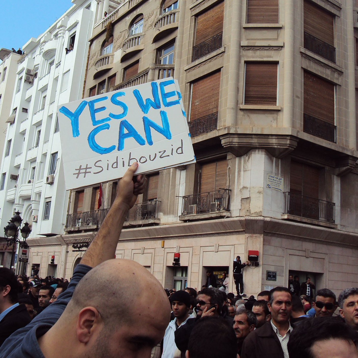 L'image montre une foule rassemblée dans une ville, probablement lors d'une manifestation. Au centre, une personne tient une pancarte avec le message "YES WE CAN" et le hashtag "#Sidibouzid". Les bâtiments en arrière-plan suggèrent un environnement urbain. L'atmosphère semble être celle d'un mouvement populaire, avec des participants engagés et déterminés.