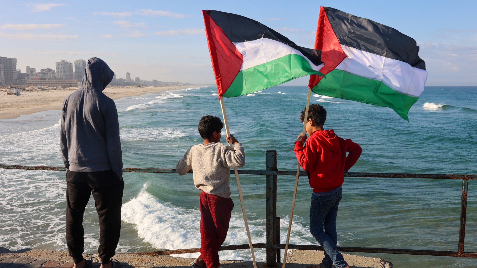 Nell'immagine, tre ragazzi sono in piedi su un molo che si affaccia su una spiaggia e sul mare. Due di loro tengono in alto delle bandiere palestinesi, che sono di colore nero, bianco, verde e rosso. Il ragazzo più piccolo, in una felpa grigia, sembra osservare attentamente l'orizzonte. La scena è illuminata da una luce naturale, con il cielo blu e alcune nuvole sparse. Le onde del mare si infrangono dolcemente sulla riva, creando un'atmosfera tranquilla. Gli altri due ragazzi indossano abbigliamento casual e sembrano condividere un momento di riflessione insieme.